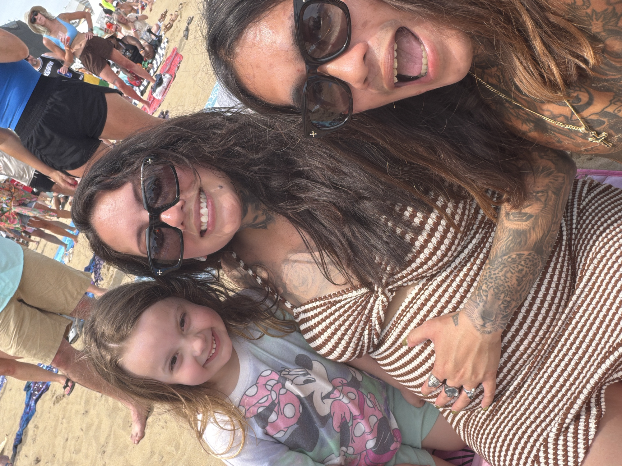 This image captures a group of people at a crowded beach, likely during a daytime event. In the foreground, three individuals are posing for a selfie. On the left, a young girl with light brown hair smiles widely, wearing a white t-shirt with a Minnie Mouse graphic and light blue shorts. Next to her, a woman with long dark brown hair and sunglasses has one hand on her stomach, wearing a brown and white striped dress. Her arm features a tattoo of a leopard. To the right of the woman, another person with long dark brown hair and sunglasses has their mouth open in an excited expression, wearing a gold chain with a cross pendant. Their torso is heavily tattooed. The background shows a sandy beach with numerous people in various casual beach attire, some sitting on towels and blankets. The presence of a large crowd suggests a public gathering or festival, common in a tourist destination like Virginia Beach, United States of America, known for its extensive shoreline and summer events. The overall atmosphere appears lively and social.