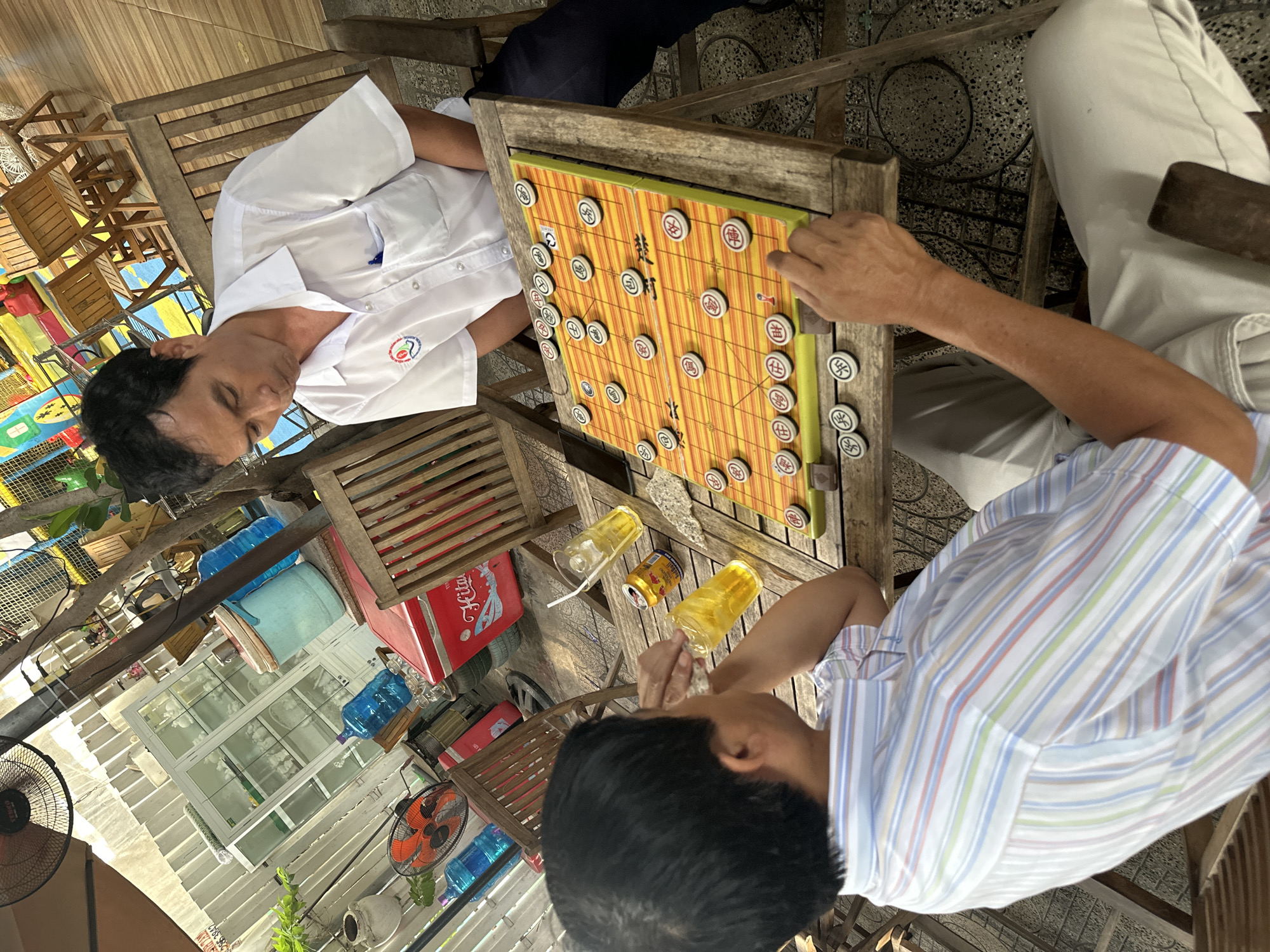 In an outdoor seating area in Nha Trang Ward, Vietnam, two men are engaged in a game of Chinese chess. The man on the right, wearing a white polo shirt with a logo on the left breast pocket and dark trousers, is looking down at the chessboard. The man on the left, wearing a striped short-sleeved shirt and light-colored trousers, is partially visible from the back and is reaching towards the chessboard. The chessboard itself is orange with yellow lines and features Chinese characters on the pieces. Two glasses of amber liquid, one with a straw, and a red can are placed on the wooden table next to the chessboard. The background shows a rustic outdoor setting with wooden chairs, a fan, a white cabinet, and red coolers.