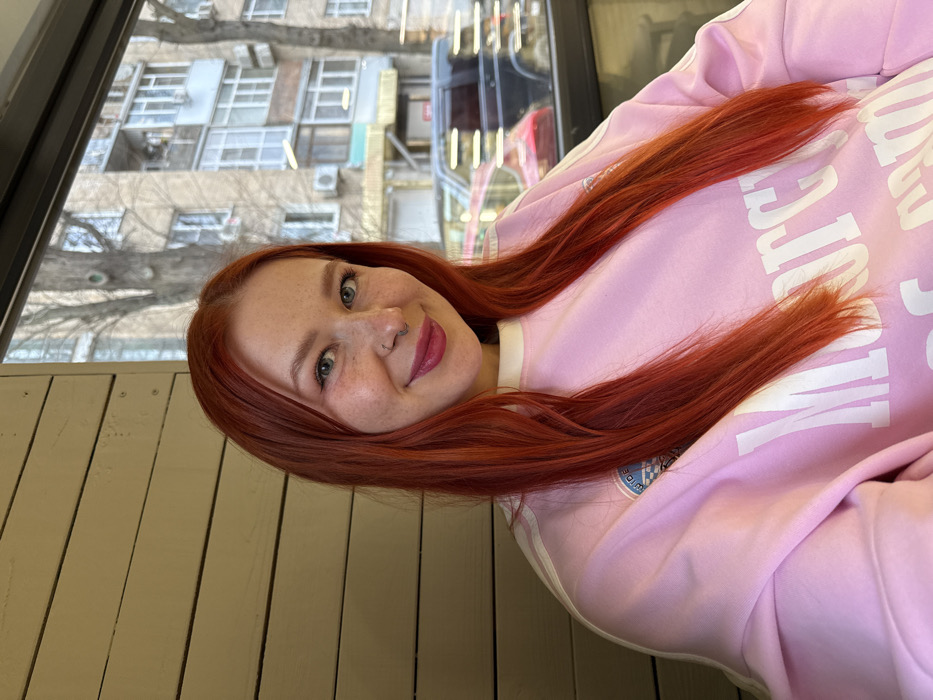 A female individual is depicted in a medium-shot, smiling and looking directly at the camera from an indoor location. She possesses long, straight, bright red hair parted in the center, blue eyes, a fair complexion with freckles, and a small silver septum piercing. Her lips are colored with pink lipstick. She wears a light pink long-sleeved sweatshirt featuring white stripes on the shoulders and sleeves, a checkered patch on the left upper arm, and partially visible white text on the chest commencing with "Mor...". The immediate interior environment includes a light-colored, horizontal wooden paneled wall to the left. To her right, a large window reveals an urban street scene in Rostov-on-Don, Russia. The exterior visible through the window features multi-story residential buildings with various windows and balconies, bare deciduous trees, and parked vehicles including a dark SUV and the reflection of a red car. The outdoor lighting is diffused, indicating an overcast condition. The indoor setting appears to be a cafe or similar establishment with a street-facing view.