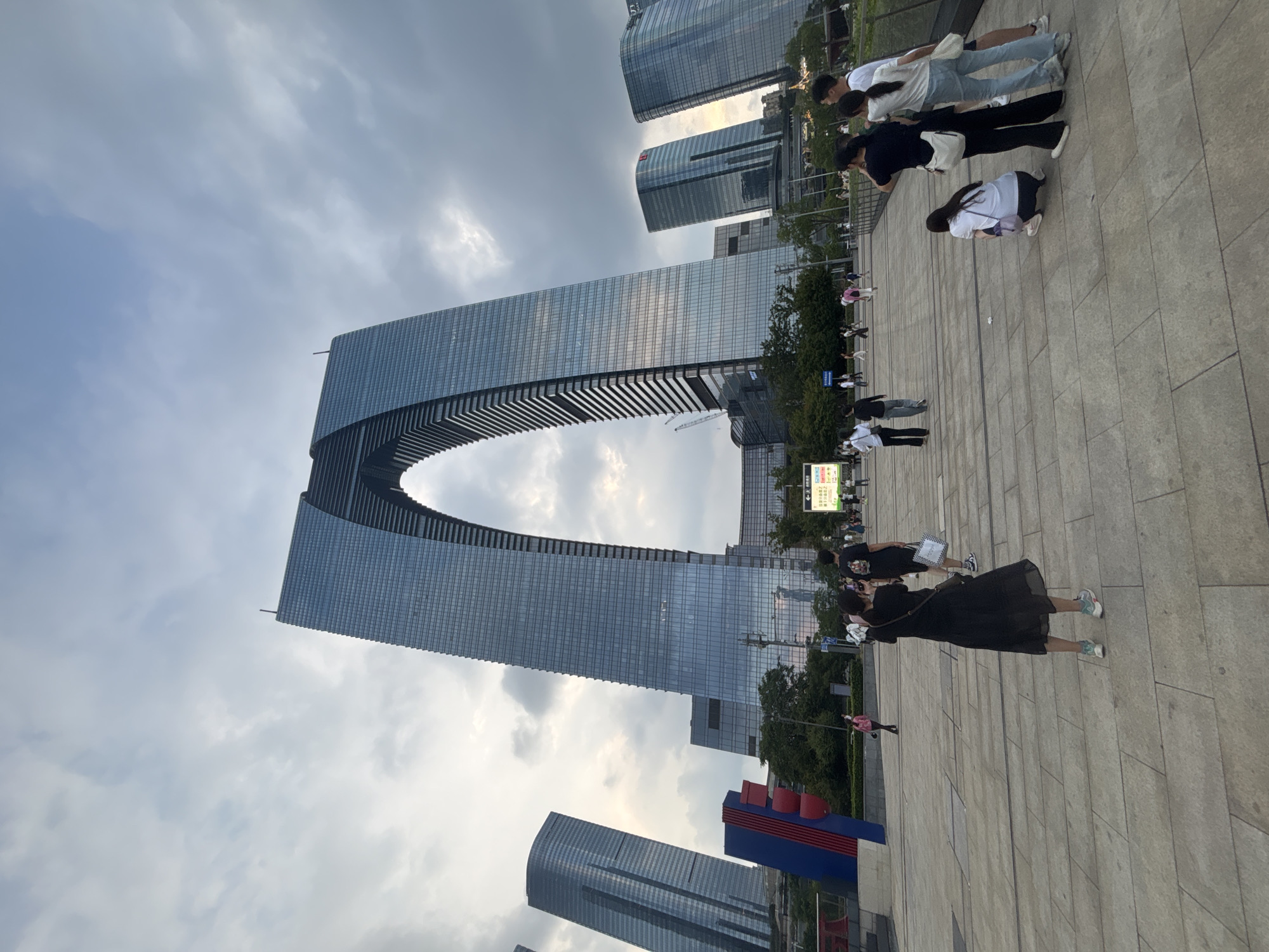 The image captures the Suzhou Landmark Building, also known as the Gate of the Orient, in Suzhou Industrial Park, China. The striking arch-shaped skyscraper dominates the skyline, its glass facade reflecting the overcast sky. In the foreground, a group of tourists are seen taking photographs and observing the architecture. The plaza area is paved with large, light-colored stone tiles, and a few trees and other modern buildings are visible in the background. The scene appears to be a tourist destination, with people engaging in leisure activities.