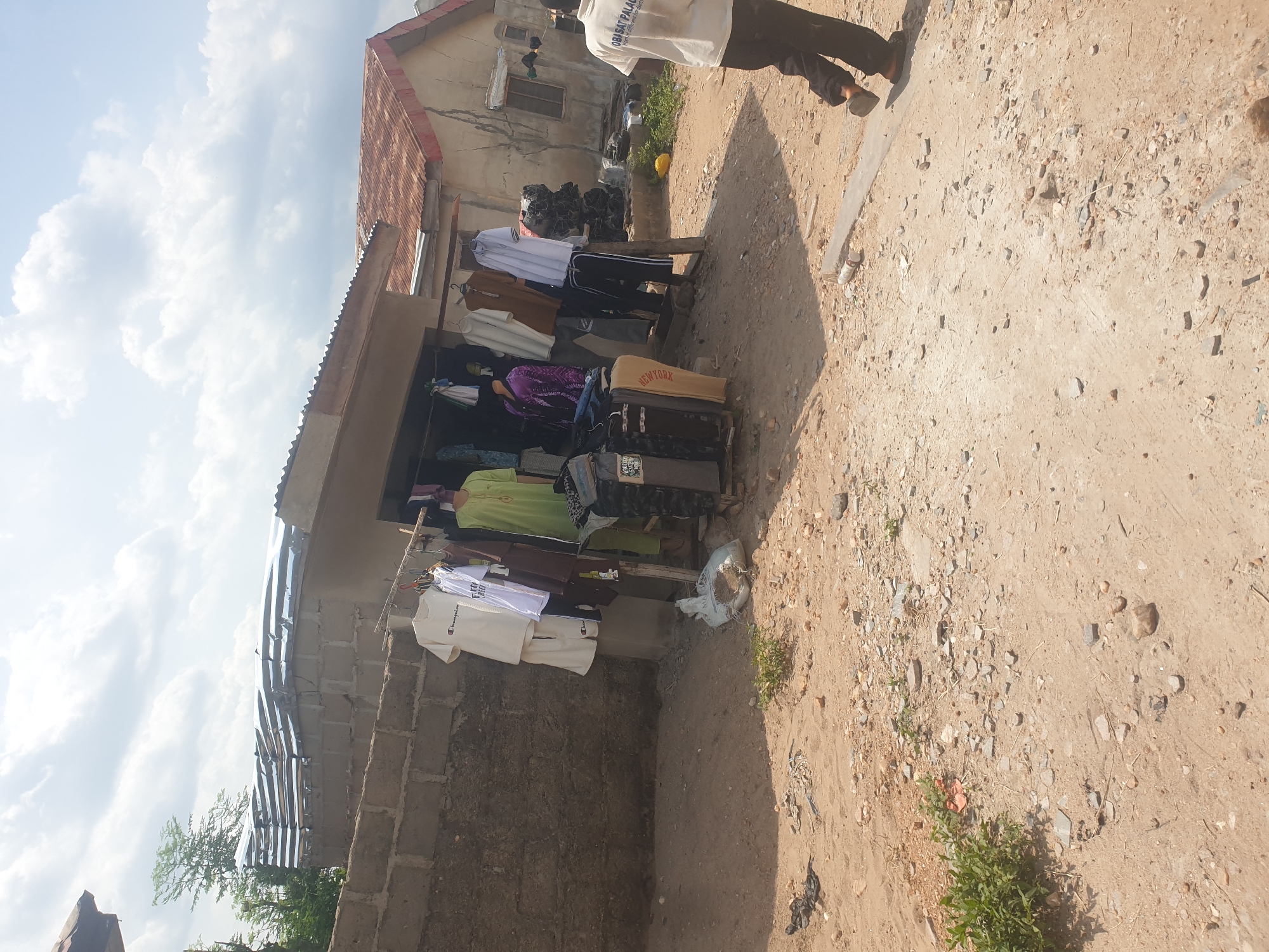 The image shows a street-side clothing shop. Clothes hang on display in front of a small building with a corrugated metal awning. A person is standing near the edge of the road, their upper body visible in the upper right corner of the frame. The road is unpaved and rocky. The sky is cloudy.