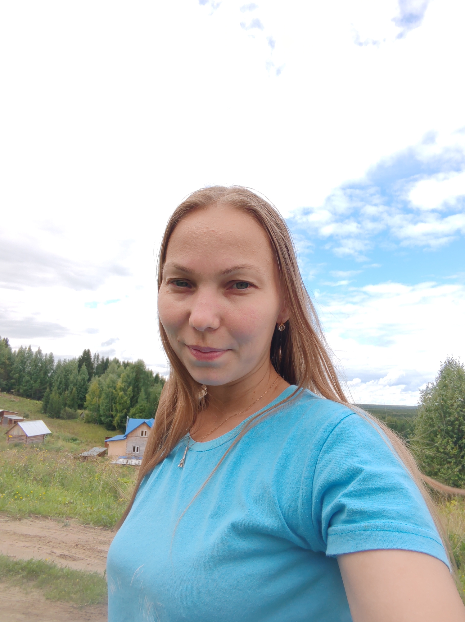 A young woman with long blonde hair, wearing a light blue t-shirt and a gold necklace, takes a selfie outdoors. She is smiling slightly and looking at the camera. Behind her, a rural landscape unfolds with a dirt path in the foreground, grassy hills, and a cluster of houses with wooden roofs. A dense forest of green trees forms the backdrop, under a partially cloudy sky with patches of blue. The location is identified as Chukhlem, Russia.