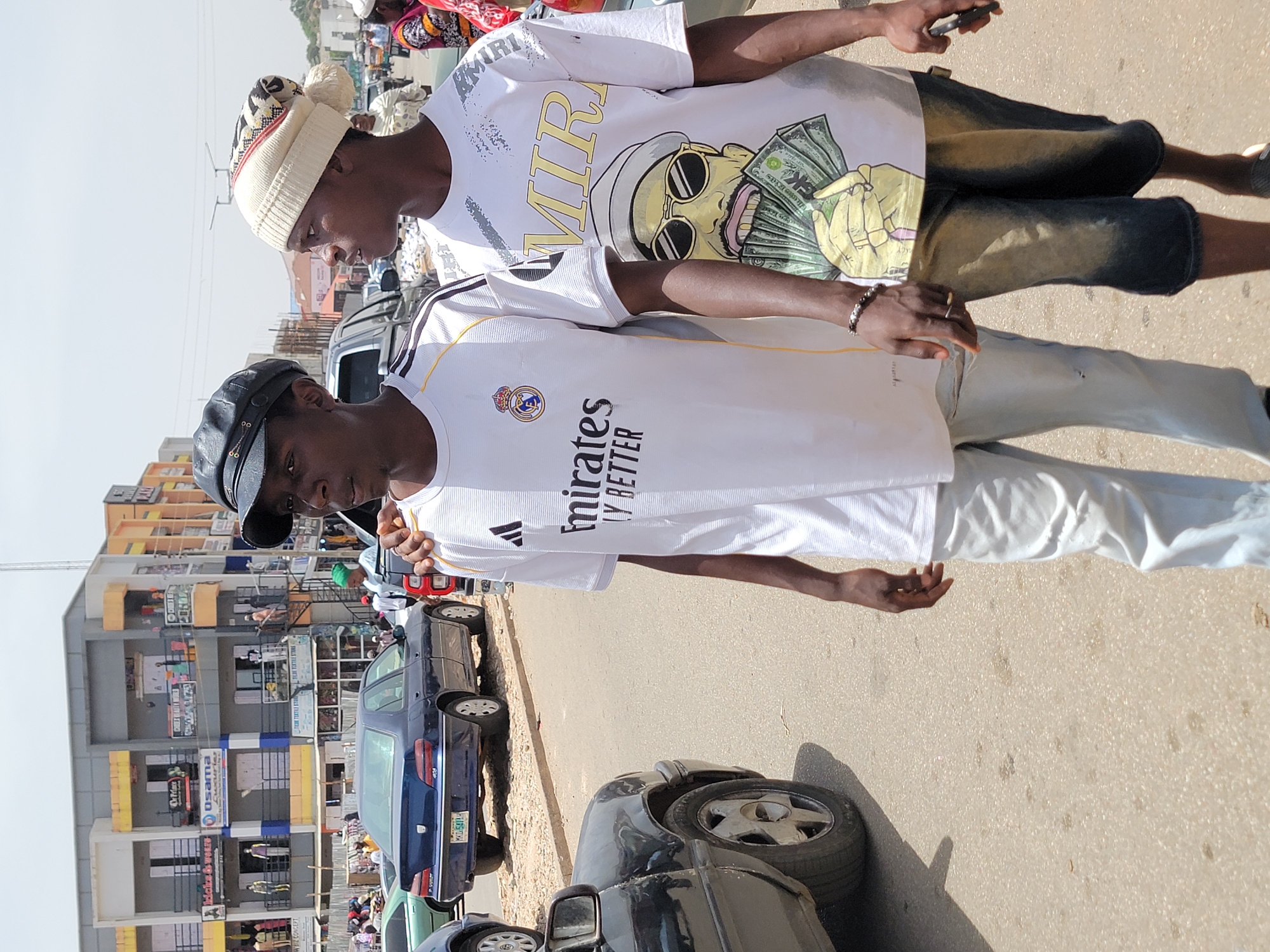 Two young men stand on a street in Jos, Nigeria, near a four-story commercial building with storefronts bearing names like "Osama" and "Whette Kids." One man wears a white t-shirt with an "AMIRI" design featuring a character with sunglasses and money, denim shorts, and a knitted cap with a pom-pom. The other man wears a white Real Madrid soccer jersey, light-colored pants, and a black cap. Cars are parked along the street.