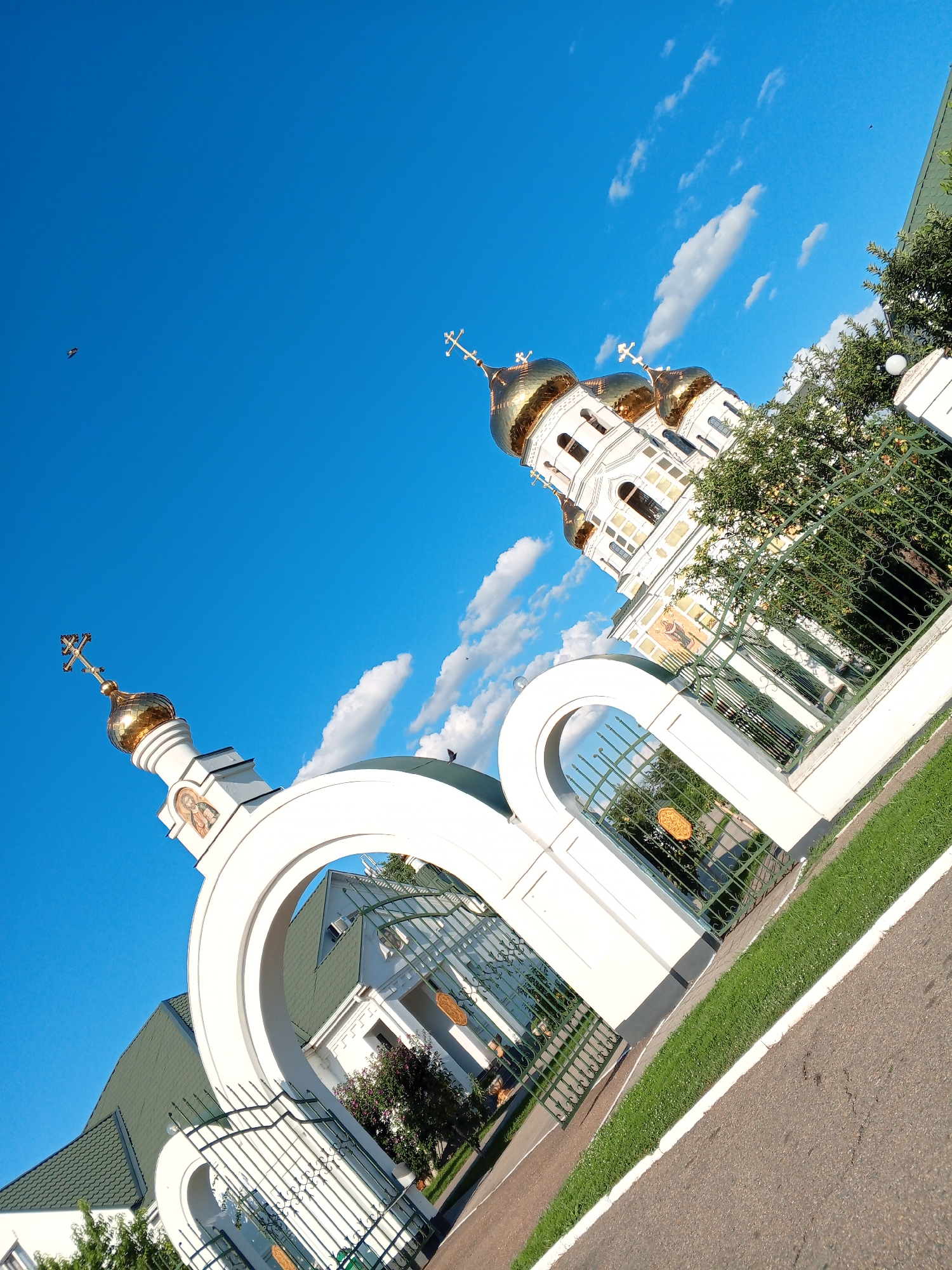 This is a photograph of the Church of the Transfiguration in Novodonetskaya, Russia, taken from a low angle, looking upwards towards the bright blue sky. The church features a white facade with golden domes and crosses. In the foreground, there is a white arched gateway with an ornate green metal fence. The grounds are covered in green grass and a paved pathway. A few wispy clouds are visible in the sky, and a bird is flying in the distance. The composition is tilted, giving a dynamic perspective.