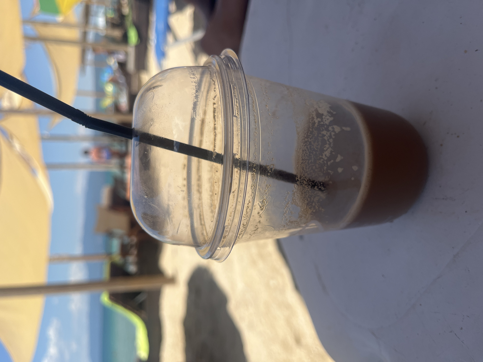 A close-up shot focuses on a nearly empty plastic cup with a domed lid and a black straw, resting on a white surface. The cup contains a small amount of brown liquid with foam residue on the sides. The background is a blurred beach scene, likely in Ofrynio Beach, Greece, under a bright, sunny sky. Beach umbrellas, lounge chairs, and the sea are visible. The image captures a moment of leisure on a warm day.