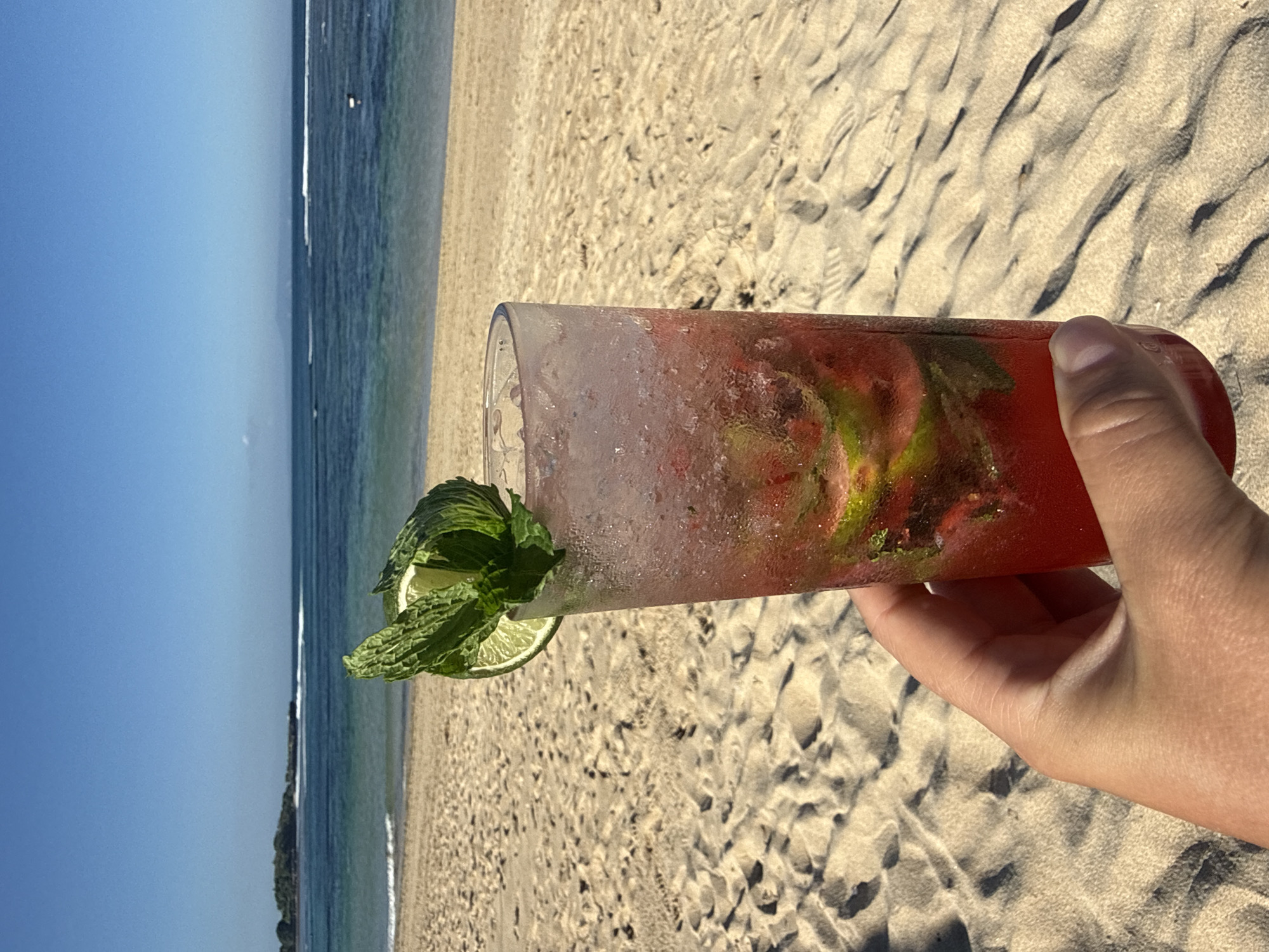A person's hand holds a tall, clear glass containing a red cocktail with ice, mint, and lime. The glass is frosted and condensation is visible. The cocktail is garnished with a lime wedge and mint leaves. The background is a sandy beach with gentle waves and the ocean stretching to the horizon under a clear blue sky. The location is likely Kuta Selatan, Indonesia, given the tropical beach setting.