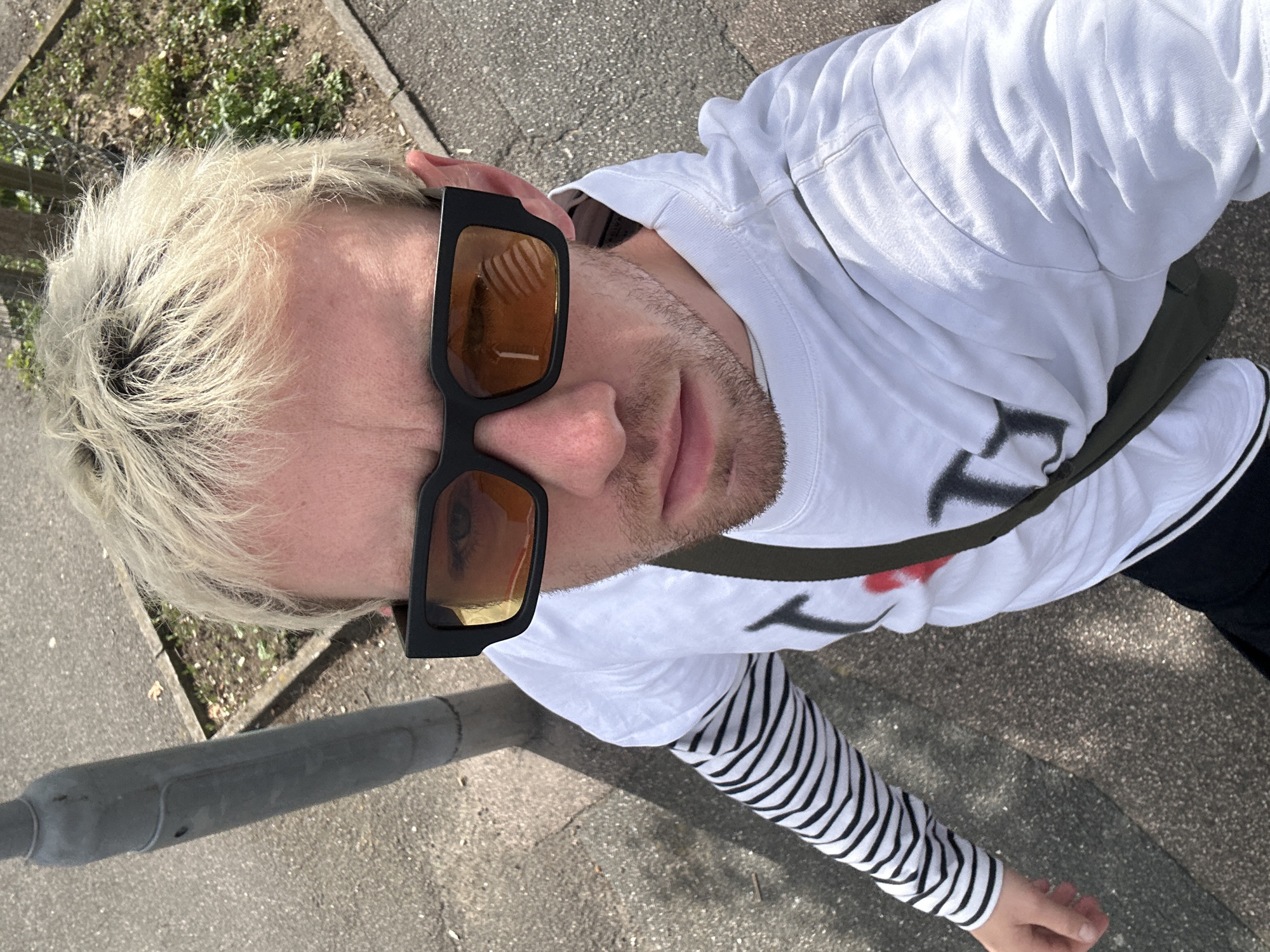 In a high-angle, self-taken photograph, a person with bleached blonde hair, wearing black-rimmed sunglasses with amber lenses, stands on a paved surface in Folkestone and Hythe District, United Kingdom. The individual is dressed in a white long-sleeved t-shirt layered over a striped long-sleeved shirt with black and white horizontal stripes. A dark green strap, possibly from a bag, is visible across the chest. The background consists of a grey concrete sidewalk, a patch of soil with sparse vegetation, and a grey metal pole, likely a lamppost. The lighting suggests it is daytime, with the sun casting shadows. The image captures a close-up view of the person's face and upper body, with the environment occupying the periphery.