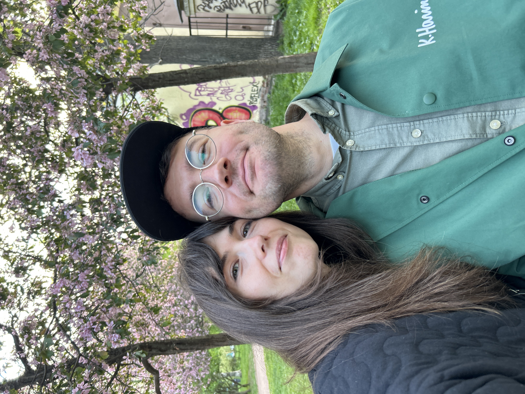 A selfie of a couple taken outdoors, likely in a park in Saint Petersburg, Russia. The man is wearing a black cap, round glasses, and a green jacket. The woman has long brown hair and is wearing a black top. Trees with pink blossoms are visible in the background, along with a graffitied wall.