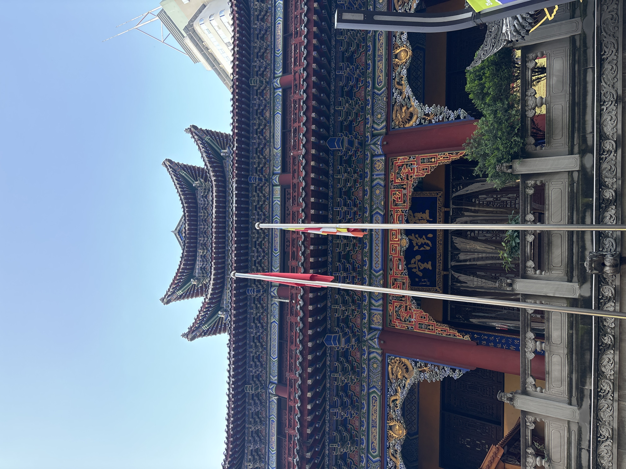 The image shows a close-up, low-angle view of a traditional Chinese temple or building. The architecture is highly ornate, with intricate details on the eaves, roof tiles, and facade. The building features multiple tiers of dark, curved roofs, adorned with colorful decorations in red, blue, green, and gold. The facade is covered in complex patterns and motifs, including what appear to be dragons or other mythical creatures.
Two flagpoles are visible in the foreground, with flags at different heights. One flag is red, and another has horizontal stripes of red, yellow, and white. The flags are made of fabric and appear to be illuminated by sunlight.
Behind the temple, a modern building with a light-colored facade and a tall antenna is visible against a clear, light blue sky. The juxtaposition of the ancient architecture with the modern building suggests an urban setting where traditional and contemporary structures coexist.
The image is taken in daylight, likely mid-morning or early afternoon, given the brightness and clear sky. The sun appears to be high, casting minimal shadows on the building. The weather seems to be clear and sunny.
There is some Chinese text visible on a blue background above an entranceway, though it is partially obscured and not clearly legible. The overall impression is one of grandeur and cultural richness, highlighting the detailed craftsmanship of traditional Chinese architecture. The scene is static, with no visible people or activity other than the flags gently blowing in the breeze, if at all. The location context provided is Yuzhong District, China.