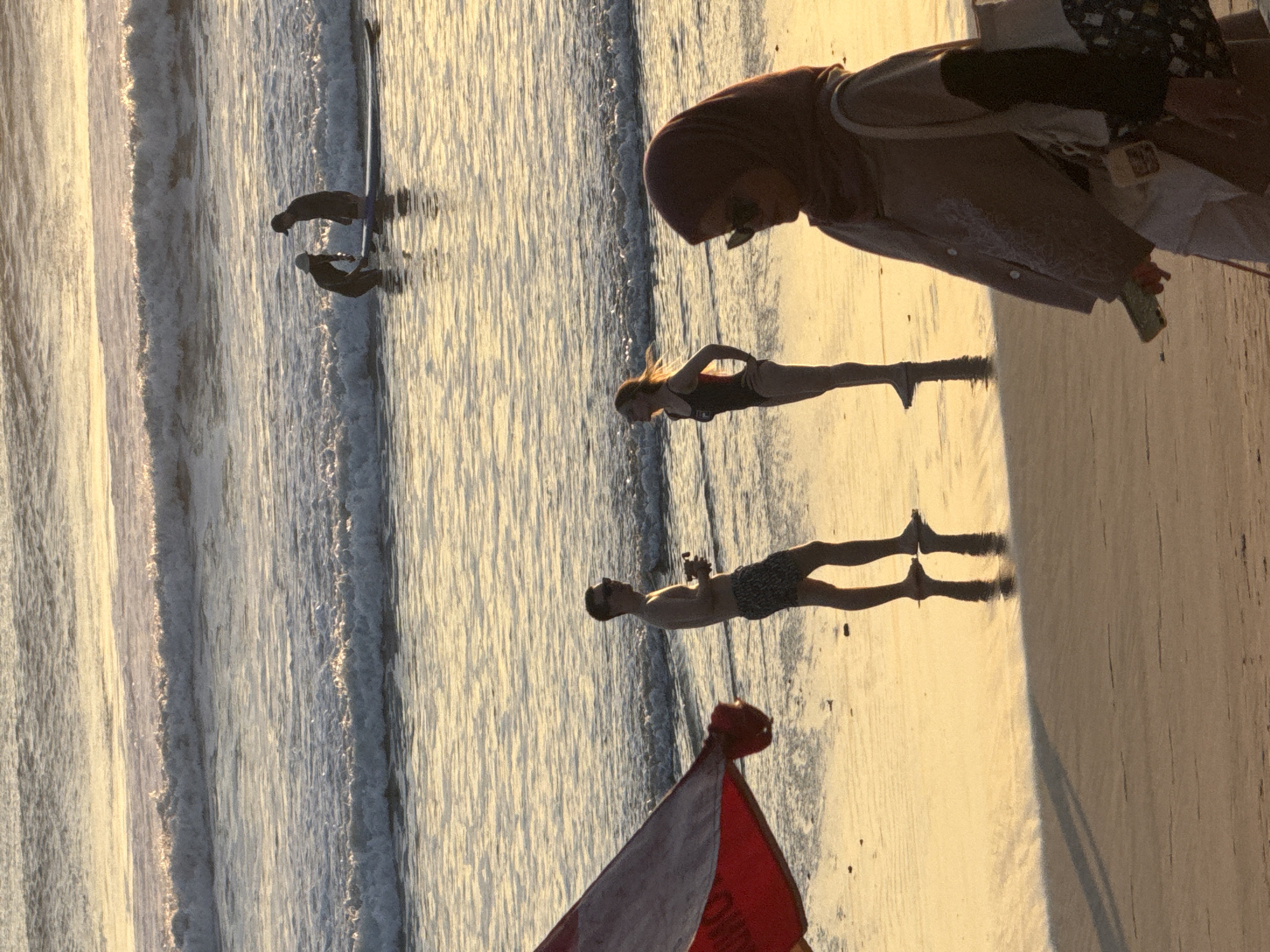 In Kuta, Indonesia, on a sandy beach during sunset, several individuals are present. In the foreground, a woman wearing a hijab and sunglasses walks across the wet sand, holding a mobile phone and a tote bag. To her left, a shirtless man in patterned swim trunks stands on the wet sand, facing away from the camera and holding a small object. Beside him, a woman in a black swimsuit stands with her hands on her hips, facing the man. In the background, further out in the surf, two figures are on a surfboard, with one appearing to be teaching the other. The ocean has gentle waves reflecting the golden light of the setting sun. A red and white beach umbrella is partially visible in the left foreground.