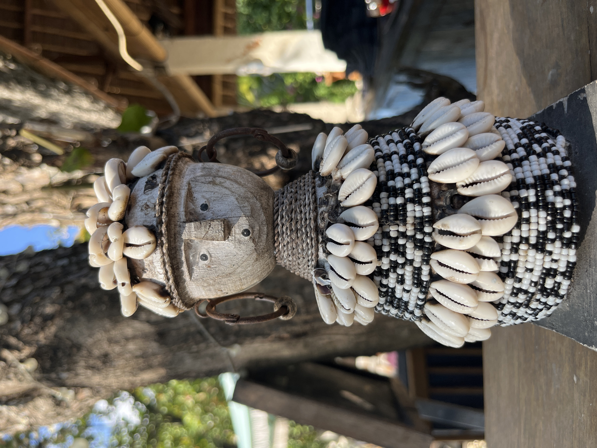 The image shows a close-up of a wooden effigy decorated with seashells and beads, situated outdoors. The effigy has a head with carved facial features, including eyes, a nose, and a mouth, and is adorned with a headdress of small white shells. The neck is wrapped in twine, and the body is conical, covered in alternating bands of black and white beads, with additional rows of shells encircling it. Two circular, rusty-colored earrings are attached to the sides of the head. The effigy is placed on a wooden surface, likely a table, with a blurred background that includes a large tree trunk, greenery, and what appears to be part of a thatched-roof structure, suggesting a tropical outdoor setting. The lighting indicates it is daytime, with sunlight casting shadows. This scene is consistent with a display of local crafts or art found in Kuta Selatan, Indonesia, possibly at a market, shop, or a cultural venue.