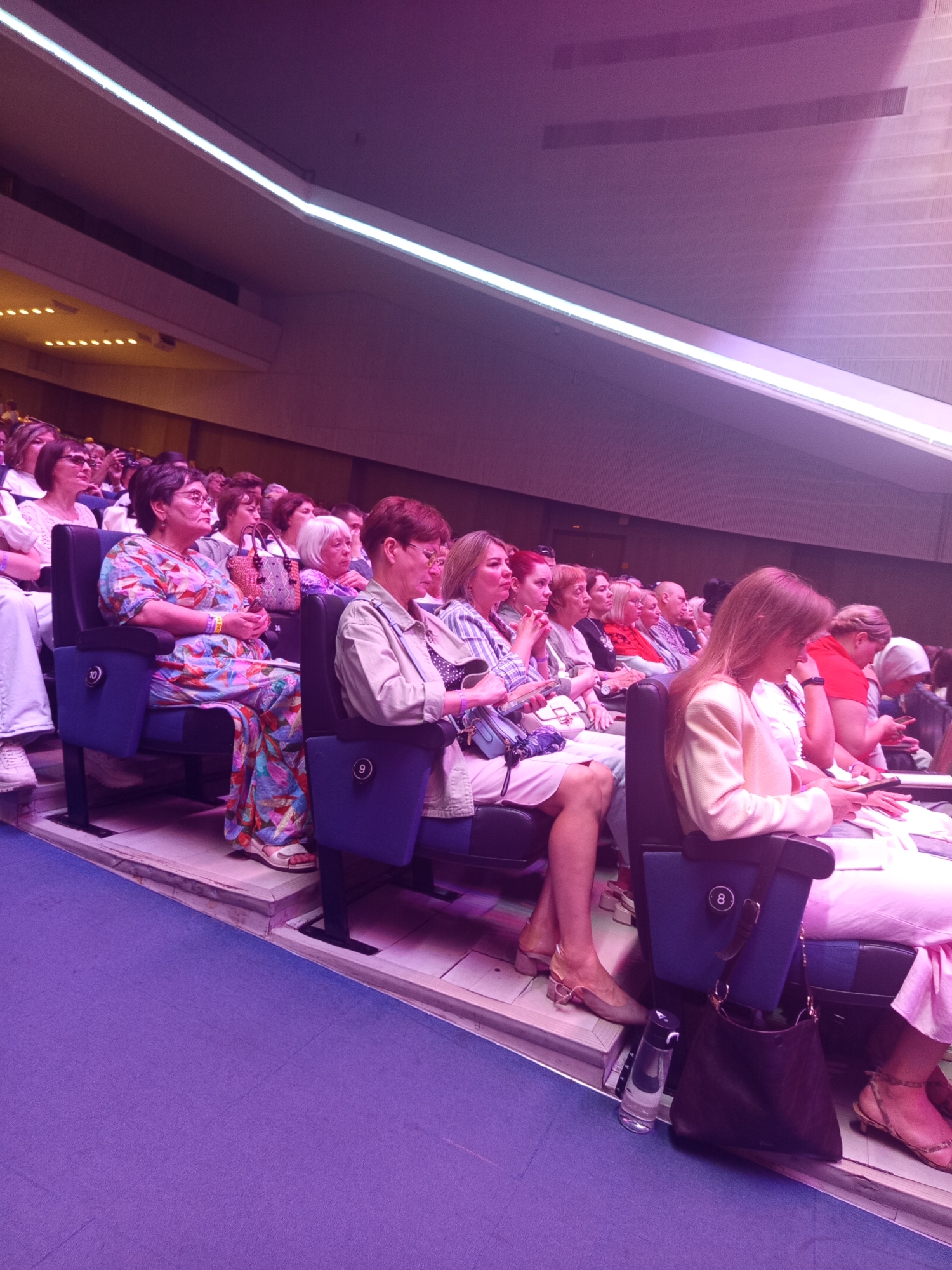 The image shows an audience seated in an auditorium. The seats are blue with a number 8 visible on the armrest of one seat. The audience members are predominantly women, dressed in casual and formal attire. Some are looking forward, presumably at a stage, while others are engaged in conversation or looking at their phones. The lighting in the auditorium is dim, with neon lights illuminating the ceiling. The floor is covered in a purple carpet. The scene appears to be a cultural event, possibly a concert or a play, taking place in Saint Petersburg, Russia, given the mention of its location in the prompt.