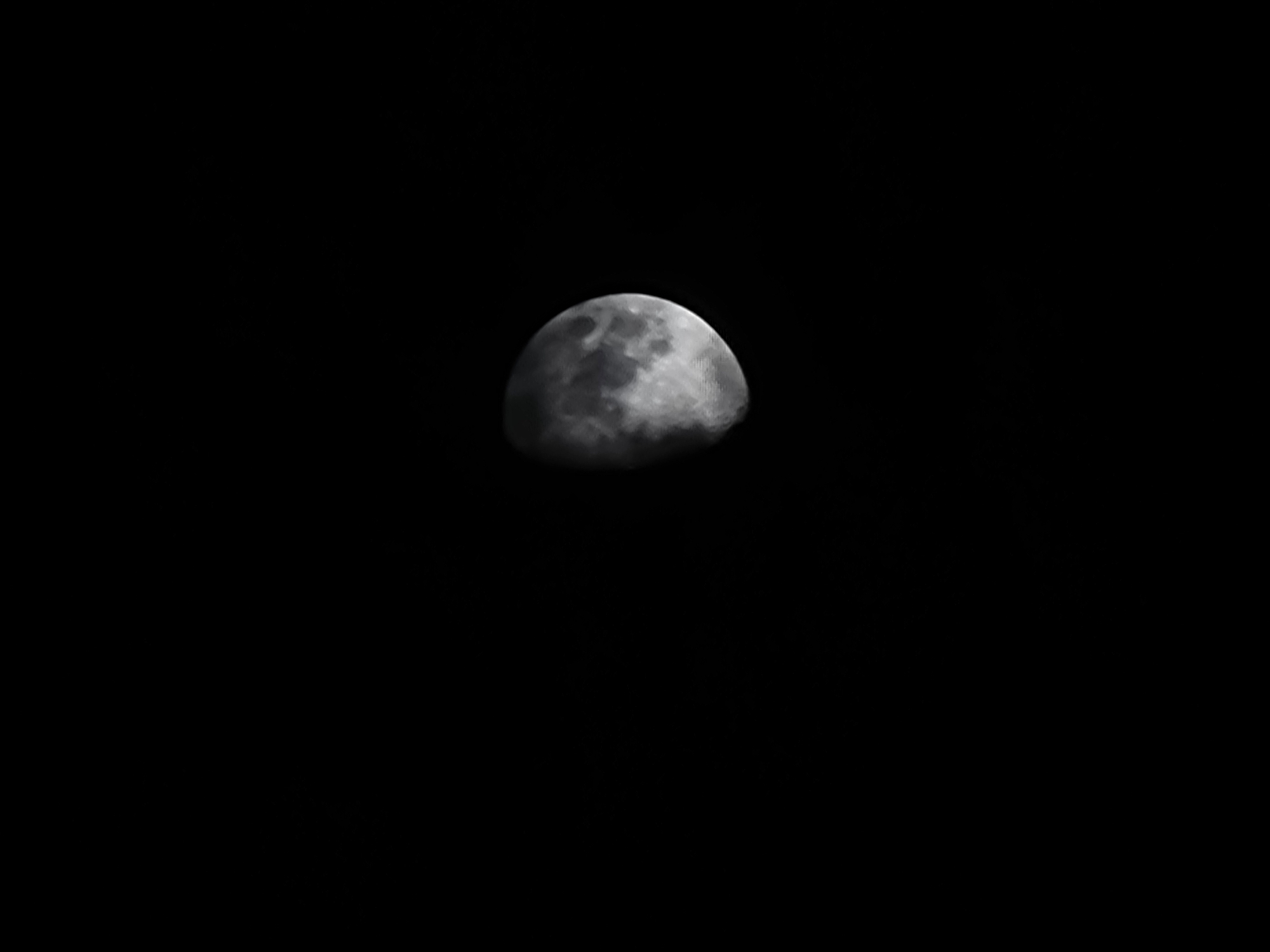 The image shows a portion of the moon against a black background. The moon is partially illuminated, with the unlit portion on the left and the lit portion on the right. The surface of the moon shows craters and maria, which are large, dark plains. The lighting suggests it is likely late evening or early morning, with the sun's rays hitting the moon from the right. No specific location in Quarteira, Portugal, can be identified from this image alone, as it depicts a celestial body visible from anywhere on Earth. The image is a close-up photograph of the moon.