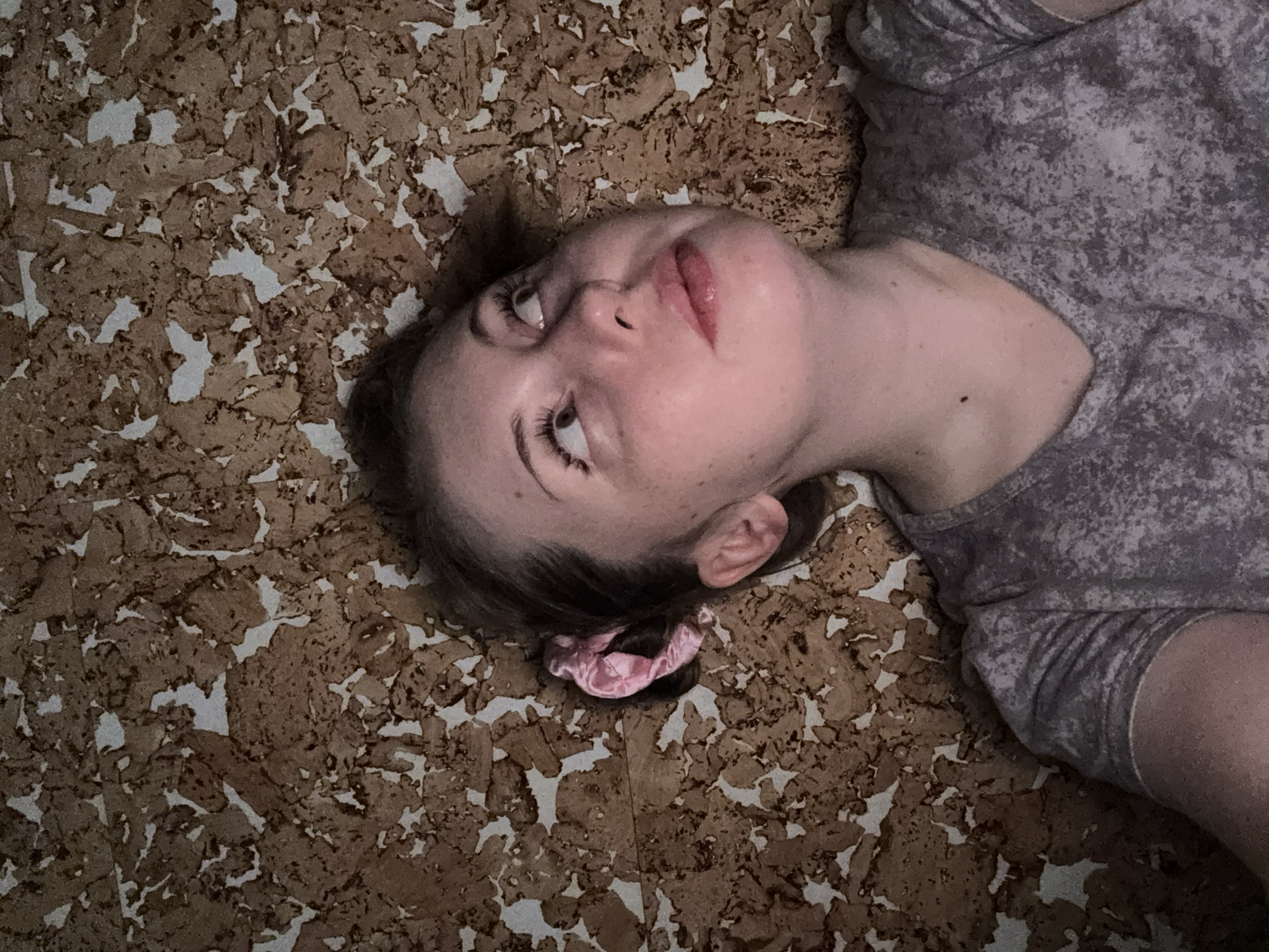 The image captures a close-up, angled view of a young woman lying on a textured surface. Her head is positioned towards the left of the frame, with her body extending to the right. She looks upwards and slightly to the right, her eyes open and her lips slightly parted. Her dark hair is pulled back, with a pink scrunchie visible behind her head on the textured background. Her skin is fair, showing some freckles or moles on her face and a small dark mole on her visible shoulder.
She wears a grey short-sleeved top or t-shirt with a mottled, light and dark grey pattern.
The background is a flat surface covered in an irregular, fragmented pattern, reminiscent of cork board or a similar composite material. It features numerous pieces of varying shades of brown and dark brown, interspersed with areas of lighter, off-white or light grey, creating a mosaic-like texture. This patterned surface fills the space around her, suggesting she is lying directly upon it.
The lighting appears dim and diffused, contributing to a soft, quiet atmosphere. There is no specific activity depicted other than her relaxed, perhaps contemplative, pose. No text is visible within the image.