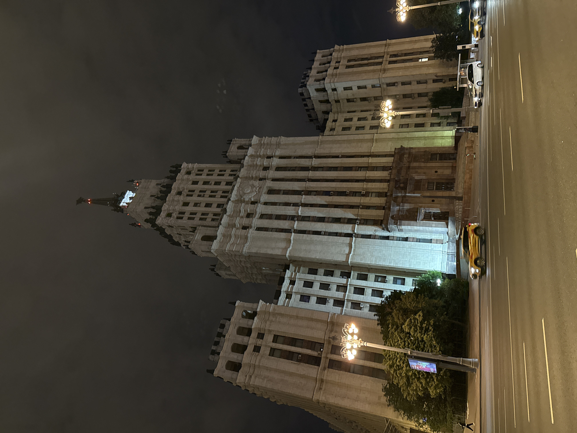 The image captures the Stalinist skyscraper, the Ministry of Foreign Affairs building, in Moscow, Russia, at night. The imposing structure dominates the frame, illuminated by streetlights and interior lighting, with the dark, cloudy sky providing a dramatic backdrop. In the foreground, a multi-lane road is visible with several vehicles driving on it. A yellow SUV is the most prominent car, with a white minivan and another vehicle further down the road. A streetlamp with multiple lights stands on the left side of the road, casting a warm glow. Trees line the sidewalk behind the streetlamp, and a digital advertisement board is also present. On the sidewalk, a lone figure walks in the distance. The architecture of the building features ornate details, multiple stories, and a prominent spire topped with a star, characteristic of Stalinist era architecture. The overall scene conveys a sense of urban grandeur and nighttime activity.
