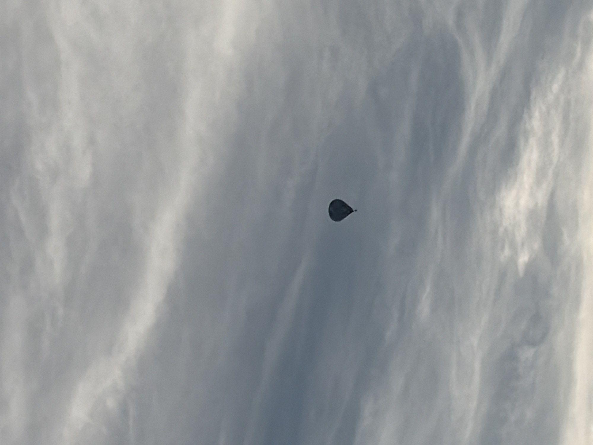 A single dark-colored hot air balloon is visible in the sky, positioned slightly to the left of the center of the frame. The balloon is a solid, dark shape against a backdrop of a cloudy sky. The clouds are varied in texture and tone, with some appearing wispy and light, while others are denser and darker, creating a sense of depth and movement. The overall lighting suggests it may be daytime, possibly overcast or nearing dawn or dusk. There are no other discernible objects or people in the image, nor any identifiable landmarks of Nitra, Slovakia.