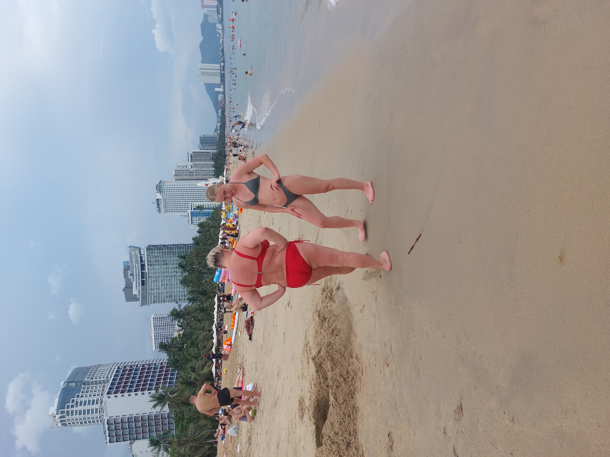 Two women are standing on a sandy beach in Nha Trang, Vietnam. The woman on the left is wearing a red bikini and is facing away from the camera. The woman on the right is wearing a dark green bikini and is facing the camera. In the background, there are several tall buildings, palm trees, and people swimming in the ocean. The sky is cloudy.