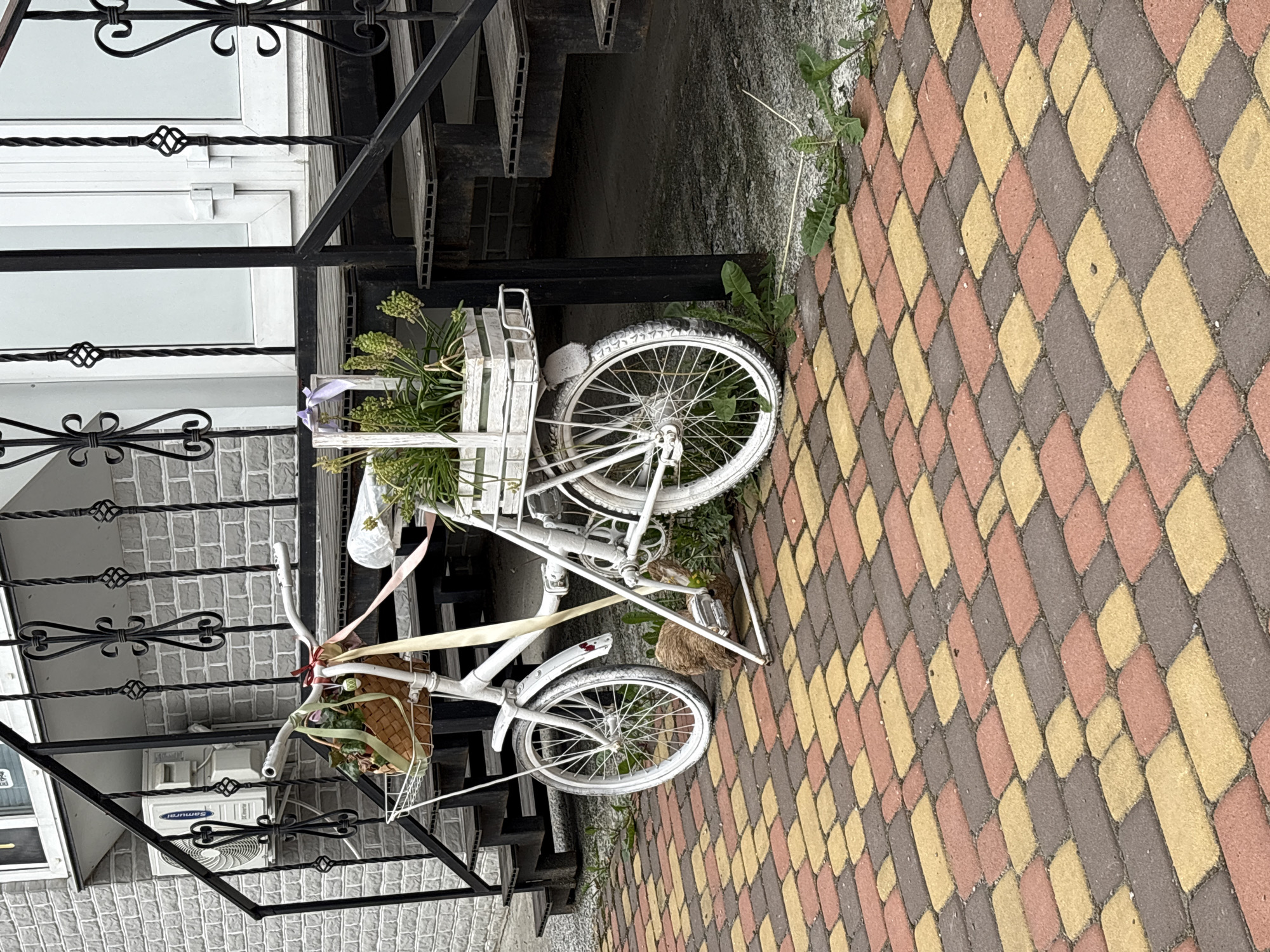 The image shows a white painted bicycle used as a decorative planter located on a brick walkway in what appears to be an urban setting in Hatne, Ukraine. The bicycle, with a wicker basket attached to the front handlebars and a wooden crate secured to the rear, is positioned adjacent to a set of stairs with a wrought iron railing. The stairs lead to a building which is partially visible. The baskets are filled with greenery and the bicycle is decorated with ribbons. The brick walkway is made up of rectangular bricks in a pattern of red, yellow, and brown. There are weeds growing near the base of the bike where it meets the brick walkway.