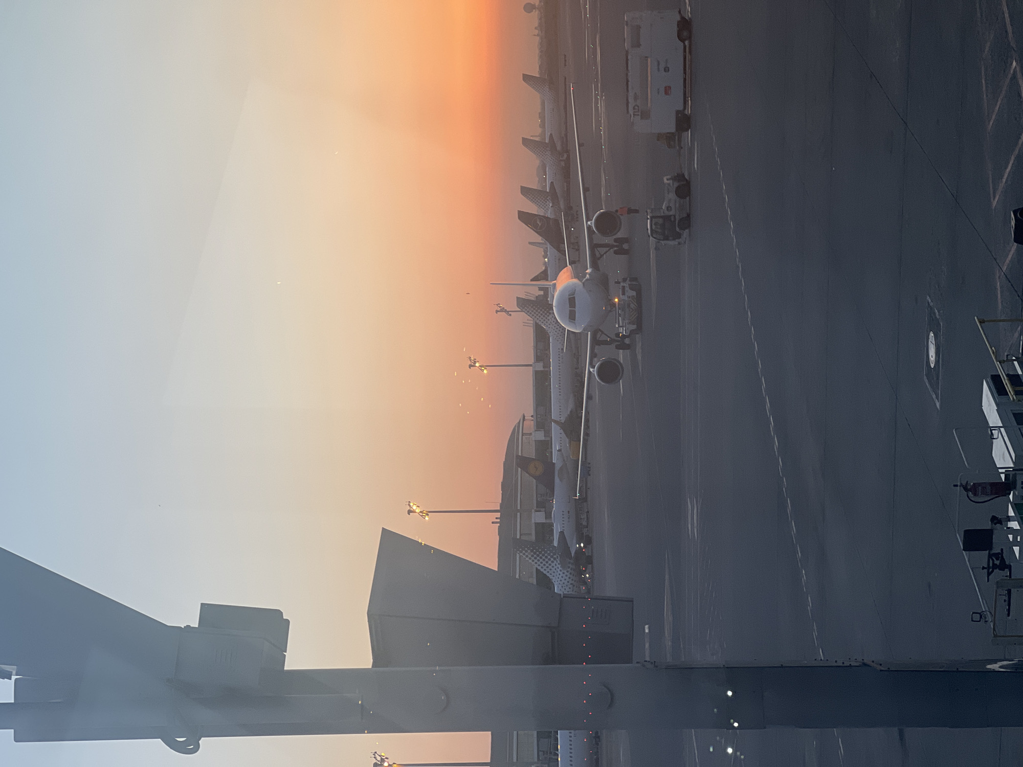 The image depicts an airport tarmac at sunset, likely at Barcelona–El Prat Airport in El Prat de Llobregat, Spain. The sky is a gradient of pale blue to warm orange, indicating the sun is low on the horizon. Several commercial airplanes are parked on the tarmac, with their tail fins visible, some featuring airline liveries. One airplane in the foreground is being prepared for departure or arrival, with a pushback tug attached to its nose. Several airport vehicles, including a white cargo truck, are present on the tarmac. The scene is viewed from an elevated position, possibly from within the airport terminal, with a large, grey structure, likely part of the jet bridge or airport building, dominating the left side of the frame. The ground is asphalt, marked with white lines and squares, indicating taxiways and parking spots. The lighting suggests it is either early morning or late evening.
