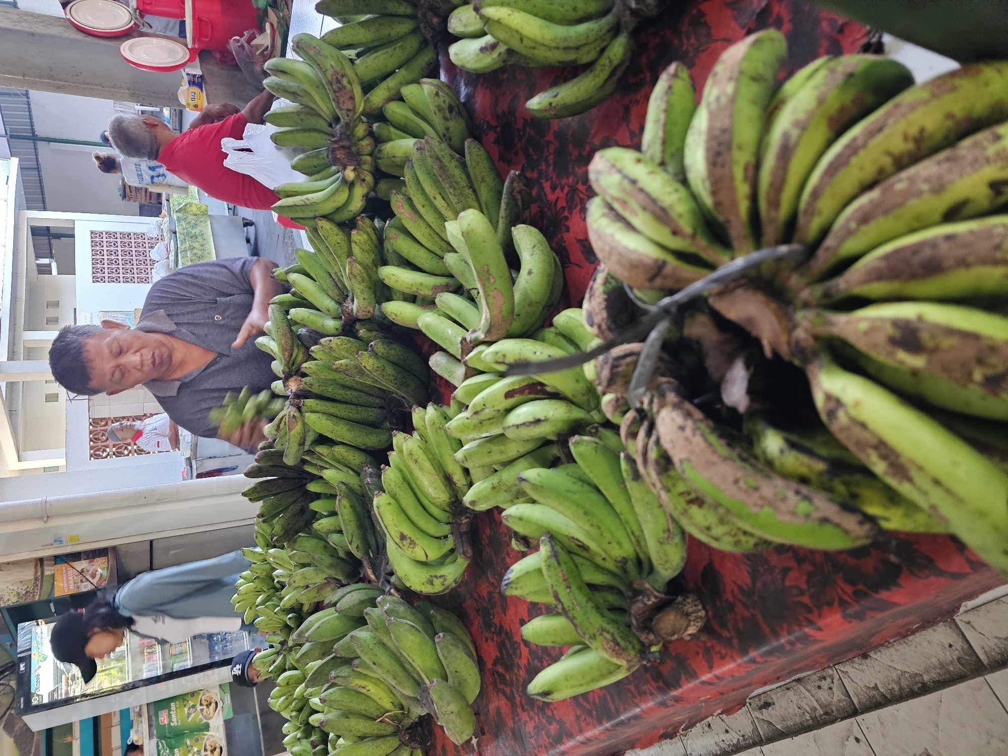 The image captures a vibrant scene at a market in Denpasar, Indonesia. An adult male with short, dark hair is selecting a bunch of green bananas from a large display. He is wearing a gray short-sleeved shirt. Numerous bunches of bananas are arranged on a red and white patterned surface. The bananas are mostly green, with some exhibiting brown spots or blemishes. In the background, another adult male in a red shirt is standing near a scale with white plates on top. Other people are visible in the periphery, including a person wearing a black cap and blue jacket. The market appears to be covered, with open-air access and visible storefronts. The floor consists of white or light-colored tiles. The scene is brightly lit.
