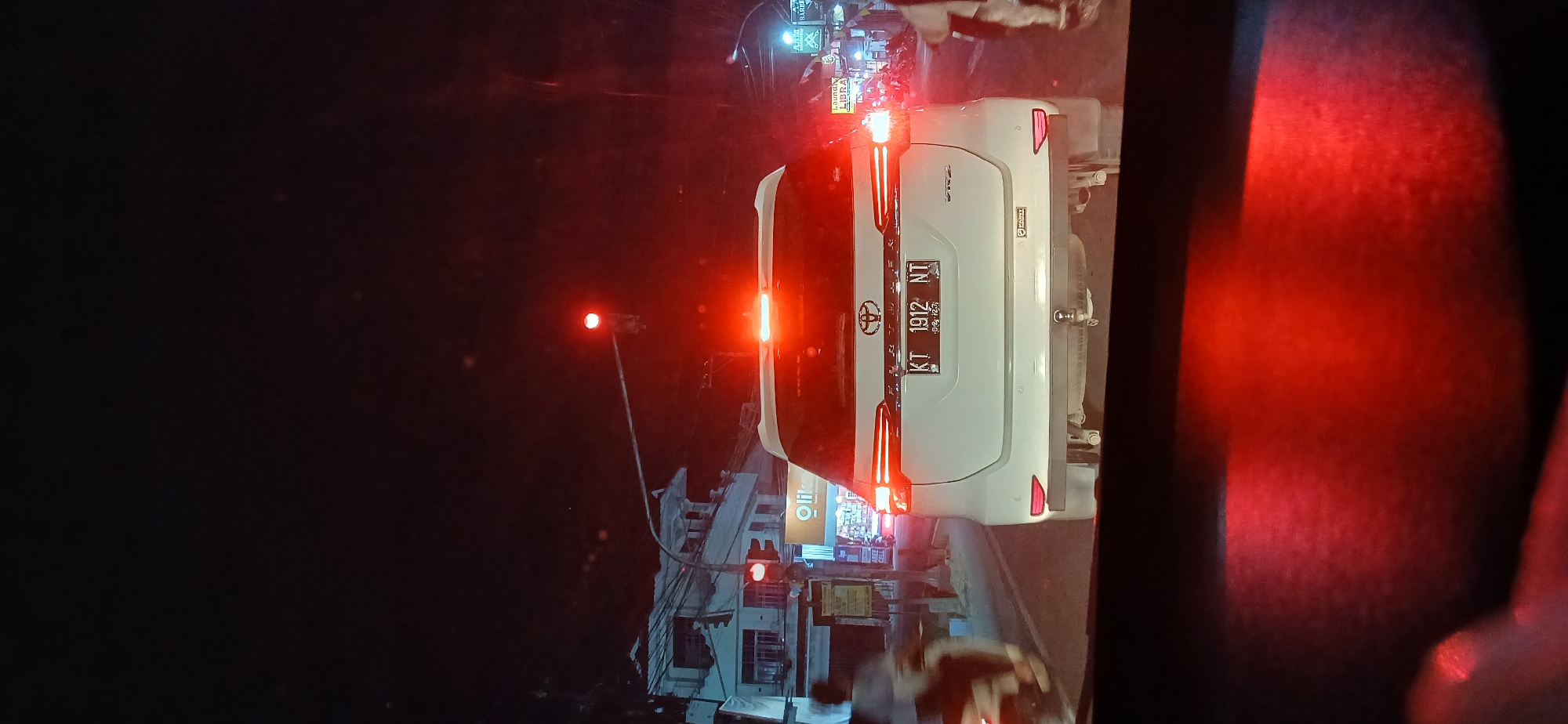 The image shows a rear view of a white Toyota Fortuner at night, stopped at a traffic light in Samarinda, Indonesia. The license plate reads "KT 1912 NT". The car is reflecting the red light of the traffic signal. In the background, there are buildings, streetlights, and other vehicles, including a motorcycle on the left side of the frame. A traffic light is visible on the right, with its red light illuminated. The scene depicts a typical urban street at night, likely at an intersection, given the presence of the traffic light and stopped vehicles. The overall impression is one of a busy city street at night, possibly in a commercial area, suggested by the presence of storefronts like "Olik" and "Laundry & Barber".