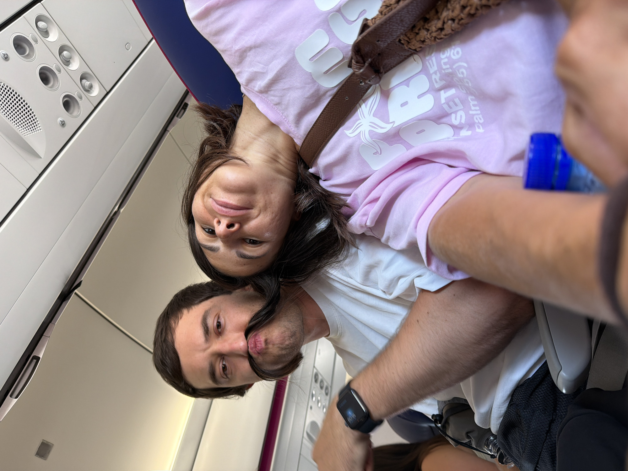 A couple is seated on an airplane, with the man in the foreground and the woman leaning her head on his shoulder. The man has a large, dark mustache made from his own hair. He is wearing a white t-shirt and a smartwatch on his left wrist. The woman is wearing a pink t-shirt and has a brown bag slung over her shoulder. The interior of the airplane cabin is visible, including overhead compartments and seats. The image appears to be a selfie taken by the couple. The location is not identifiable as Donja Cijevna, Montenegro.