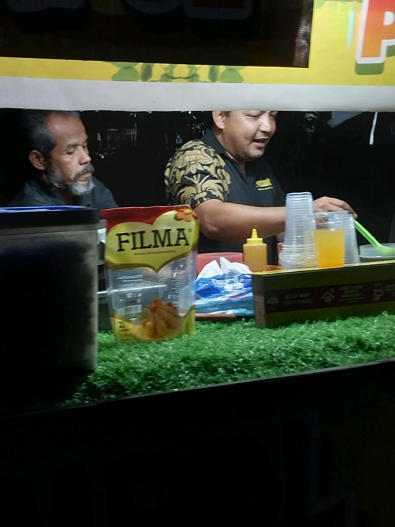 The image shows a food stall in Nglegok, Indonesia, at night. Two men are visible behind the counter. The man on the left has a gray beard and is wearing a dark shirt. The man on the right is wearing a black shirt with a patterned design on the sleeves and is preparing drinks. In front of them, there is a display of plastic cups, a bottle of orange liquid, and a package of "FILMA" branded food. The counter is covered with green artificial grass. The background is dark, with some blurry signage and lights visible.
