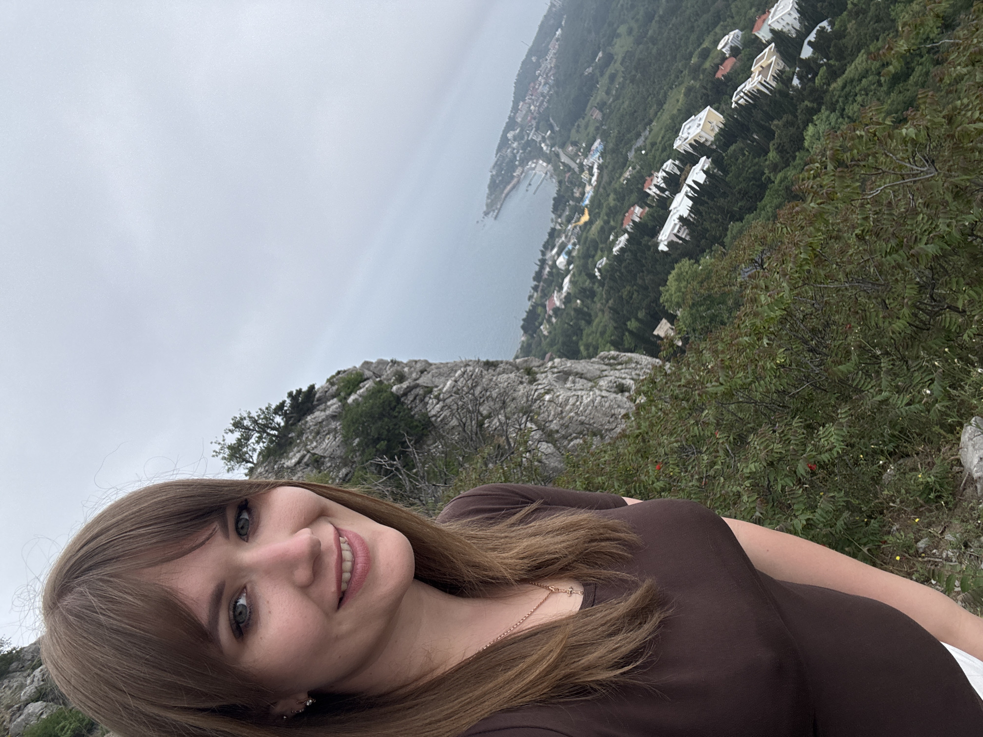 A woman stands on a rocky hillside overlooking the coastal town of Симеизский поселковый совет, Ukraine. The sea is visible in the background, along with buildings and greenery. The sky is overcast. The woman is wearing a brown top and a gold necklace. She has brown hair and is smiling.