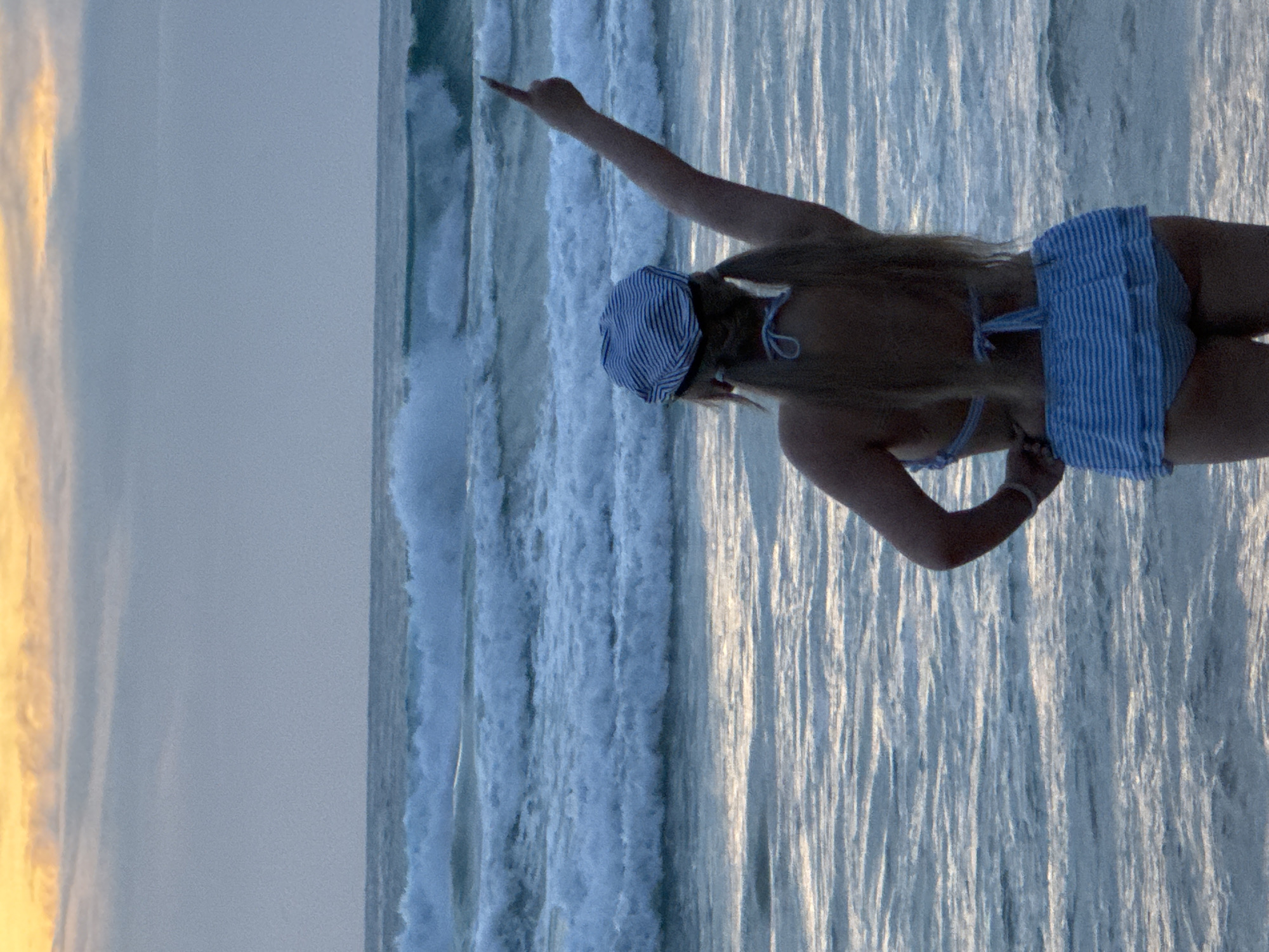 A woman wearing a blue and white striped bikini and matching hat stands on a beach in Karon, Thailand, facing away from the camera. Her right arm is raised and her index finger is pointed upwards. The ocean with waves stretches out behind her, and the sky is overcast with a hint of orange from the setting sun.