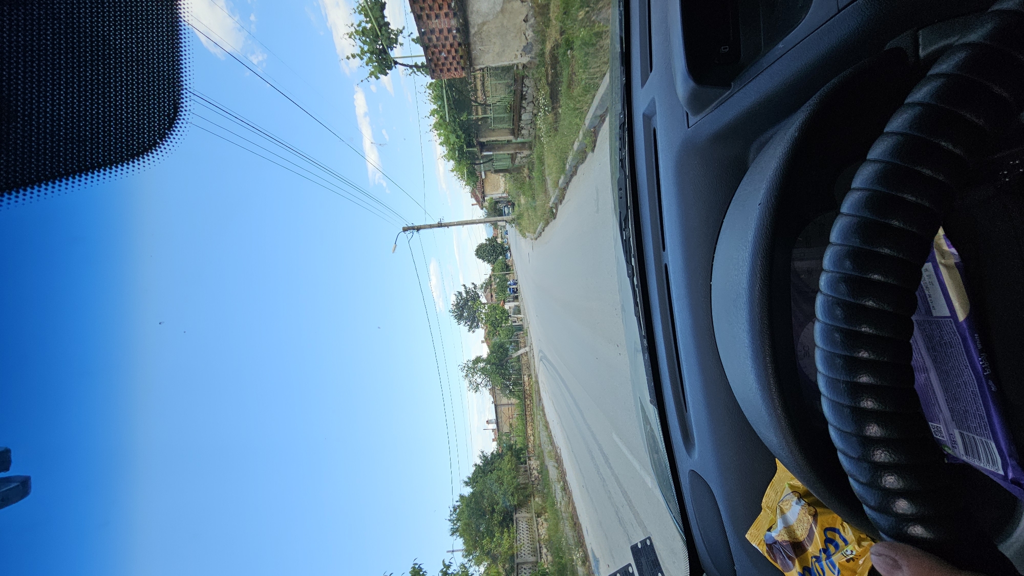 The image provides a driver's-eye view from inside a vehicle, looking out onto a narrow village road under a bright, clear blue sky with a few scattered clouds. The original image is rotated 90 degrees clockwise.
In the foreground, the dark grey dashboard and a black, ribbed steering wheel are visible on the right side of the frame. Tucked behind the steering wheel is a purple chocolate bar wrapper, partially revealing the words "Milk Chocolate" and a QR code. A yellow snack wrapper, possibly for "Lupu" branded cookies or biscuits, lies on the dashboard to the lower right. The upper left corner of the windshield shows a black, dotted frit band.
Outside the vehicle, a paved road stretches ahead, flanked by older, rustic houses on the left. These houses feature red tile roofs, worn stucco walls, and are often covered in green vines and foliage. A wooden utility pole with power lines and a street light stands prominently on the left side of the road. Abundant green trees and bushes line the street and surround the houses, suggesting a verdant, semi-rural setting consistent with Voiniagovo, Bulgaria.
The bright sunlight and clear sky indicate a sunny daytime, likely late morning or early afternoon. No people or animals are visible in the scene. The overall impression is one of a quiet, rural village road on a pleasant day.