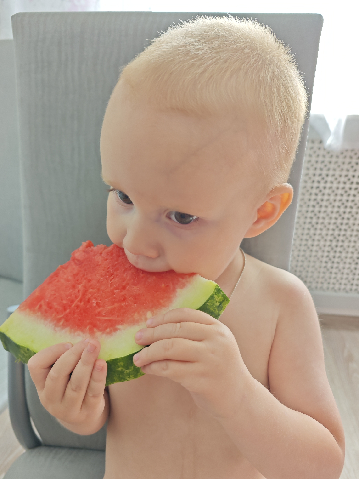 In Markovskii, Russia, a young child with light blonde hair and fair skin is sitting in a high chair, eating a slice of watermelon. The child is facing to the left, with their right hand holding the watermelon to their mouth. The watermelon is a triangular slice, showing the red flesh and green rind. The background consists of a grey chair, a wooden floor, and a patterned radiator. The lighting suggests an indoor setting, possibly a home.