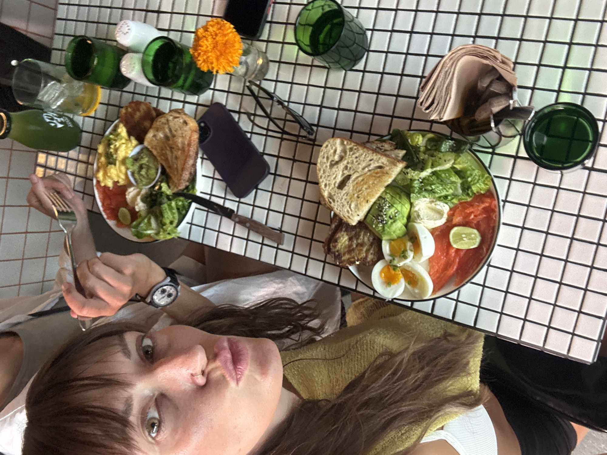 A woman, with long brown hair and bangs, is taking a selfie at a cafe. She is wearing a yellow top and has a nose ring. Her mouth is pursed. In the background, another person is visible, holding a fork. They are sitting at a table with a black and white tiled surface.
On the table, there are two plates of food. The plate closer to the viewer contains a sandwich with avocado and lettuce, sliced hard-boiled eggs, and what appears to be salmon. The other plate, further away, has scrambled eggs, avocado, toast, and tomatoes. Several green glasses, a bottle of green liquid, salt and pepper shakers, a small orange flower in a vase, a pair of tweezers, a smartphone, and a folded stack of napkins are also on the table.
The cafe appears to be in North Kuta, Indonesia. The background shows a tiled wall.