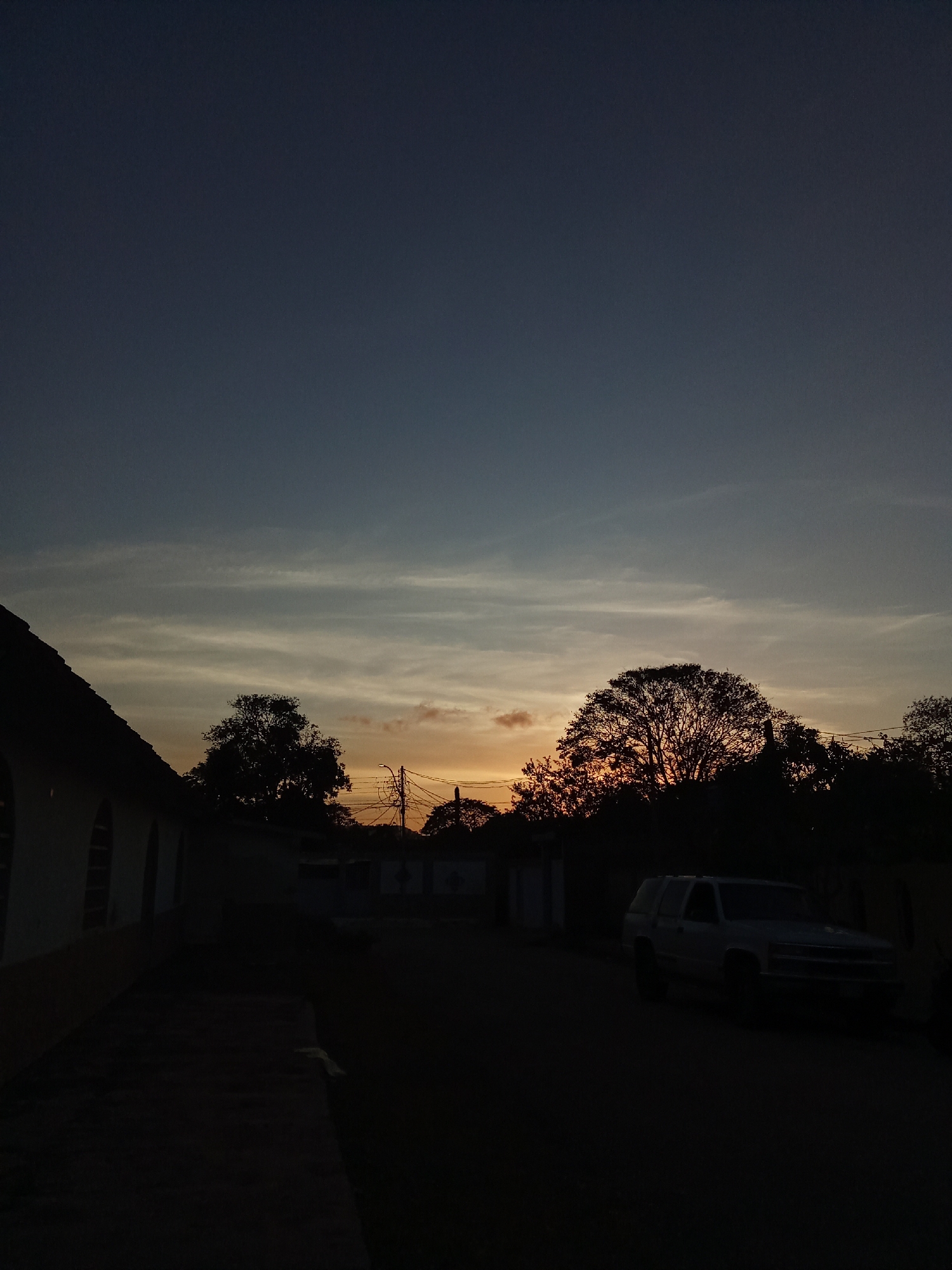 The image captures a street scene in Juan Griego, Venezuela, at dusk. The sky transitions from dark blue at the top to orange near the horizon, partially obscured by silhouetted trees. A building with a yellow stripe along its side is visible on the left. On the right side of the street, a white SUV-style vehicle is parked. Overhead power lines are visible. The street itself is dark, obscuring detail.