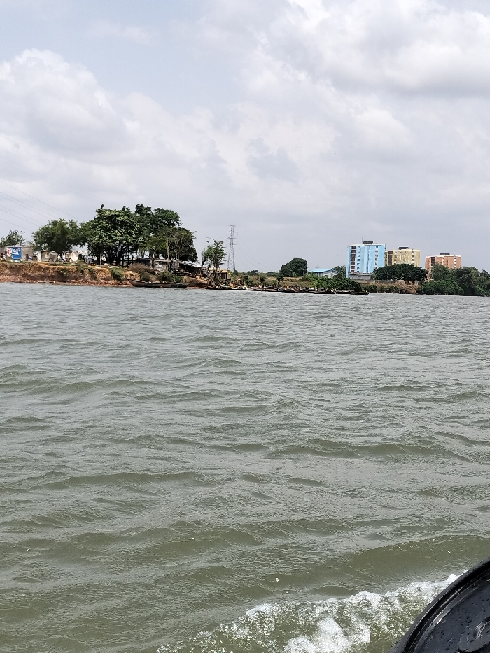 The image depicts a panoramic view of the Oworonshoki shoreline in Nigeria, observed from a body of water, likely the Lagos Lagoon. The foreground consists of dark, rippling water, with white foam and a partial black object, possibly a boat's hull or engine, visible in the bottom right, indicating the viewpoint is from a moving vessel.
The midground features a prominent earthen bank bordering the water, topped with a dense cluster of green trees. Several small, dark-colored wooden canoes are partially beached or docked along this section of the shore. Behind the trees, power lines are visible, along with a large metal lattice transmission tower. To the far left, a blue and white billboard or sign is partially obscured by foliage.
The background extends along the shoreline to the right, revealing more vegetation and three distinct multi-story buildings. From left to right, these buildings are colored light blue, yellow, and reddish-brown, suggesting residential apartments. The sky above is overcast with a mixture of grey and white clouds. No individuals are clearly discernible in the scene.