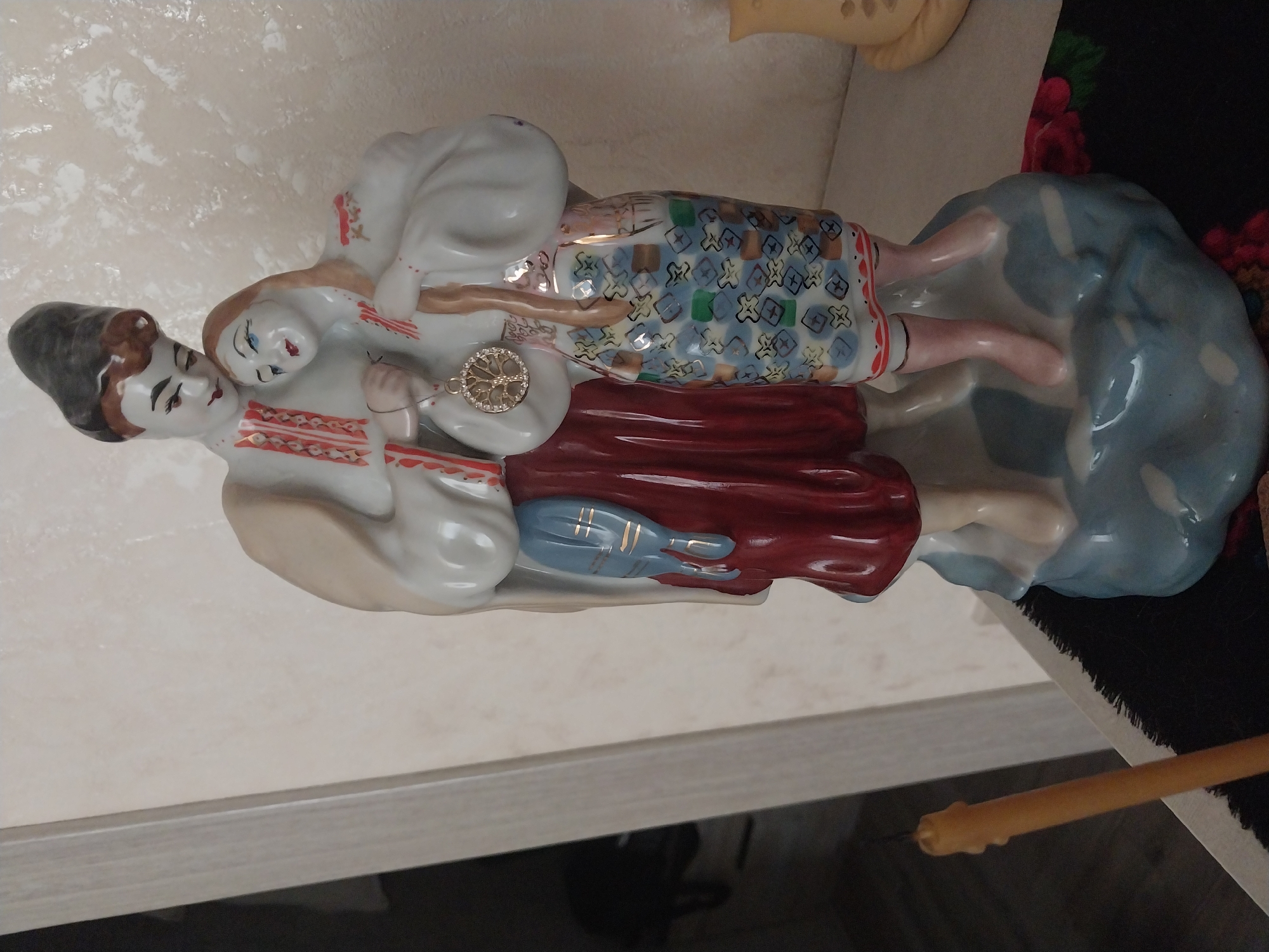 A porcelain figurine of a couple in traditional Ukrainian clothing stands on a blue, cloud-like base. The male figure wears a tall black hat, a white shirt with red embroidery, and a red skirt. The female figure has her head resting on the male's shoulder and wears a white shirt with red embroidery and a patterned skirt. A medallion hangs from a string around the male figure's neck. To the left of the figurine, a yellow candle stands upright. The background consists of a textured, light-colored wall. A dark cloth with floral patterns is visible beneath the figurine. The scene appears to be indoors, on a surface that might be a shelf or table. The lighting is soft and diffused. There is no discernible location in the city of Dnipro, Ukraine, associated with this image.
