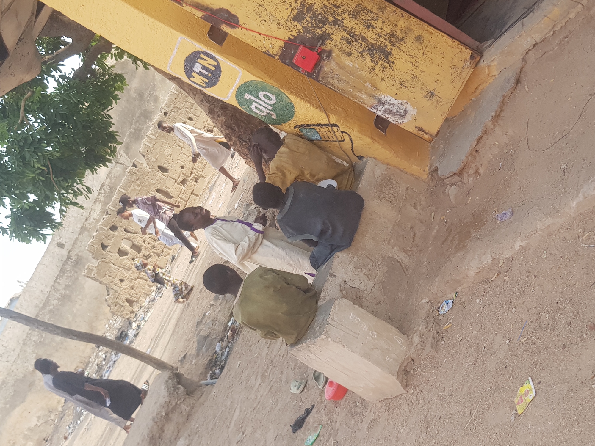 The image shows a street scene in Dutsinma B, Nigeria. Several individuals are present, some standing and walking in the background, while others are closer to the camera, seated on the ground against a yellow painted wall. This wall features logos for "MTN" and "Glo," suggesting a business or advertising. The environment is dusty and appears to be a low-income area, with simple buildings and debris scattered on the ground. The people are wearing casual clothes, including traditional robes. There is a concrete block with writing on it near the seated figures.