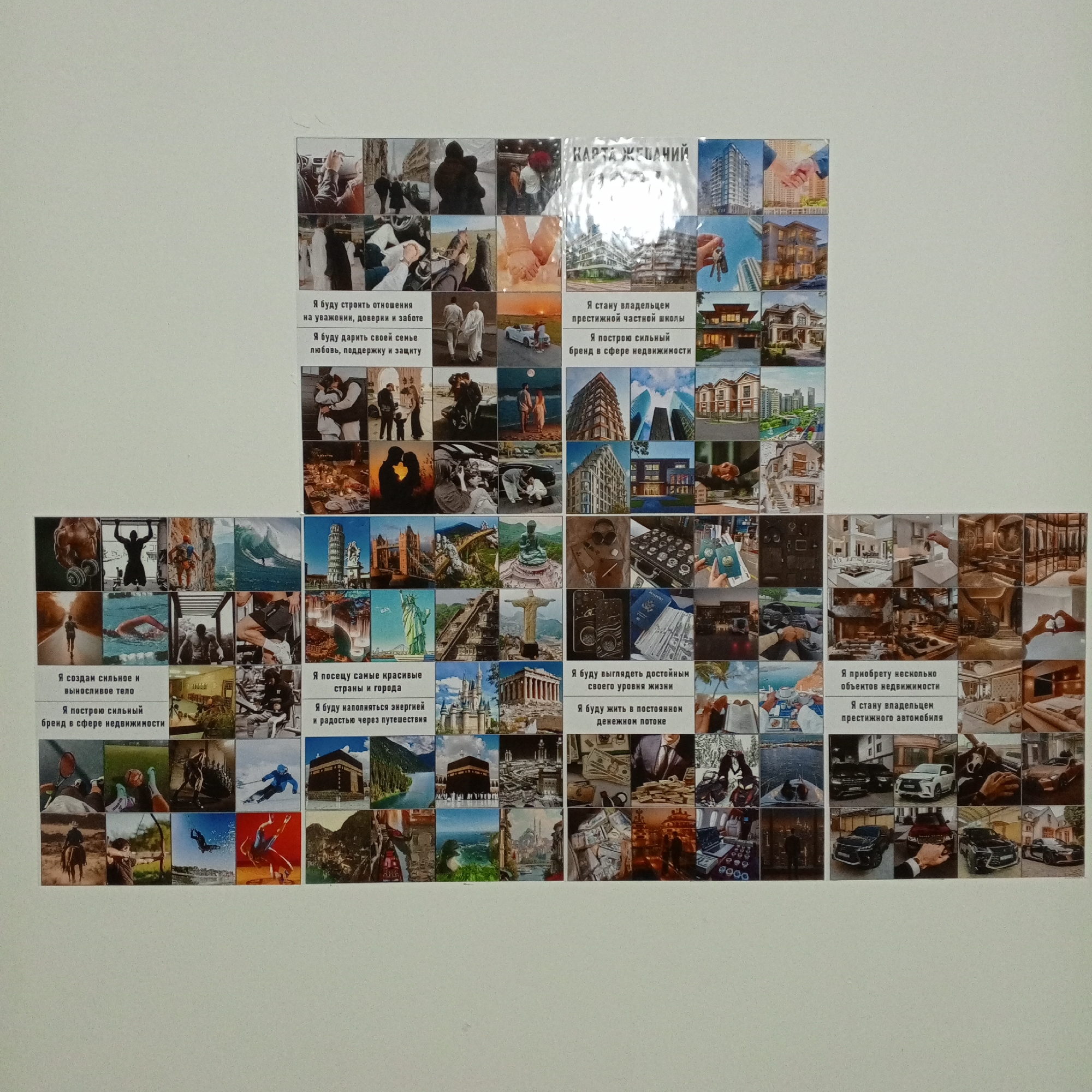 The image displays a large vision board composed of many smaller photographs and text snippets arranged on a white wall. The vision board is centered in the frame, with a significant portion of the white wall visible around it. The photographs depict a variety of aspirations, including images of successful individuals, luxurious homes, expensive cars, travel destinations, and depictions of strong family bonds and physical fitness. Some of the text, written in Cyrillic script, outlines goals such as building strong relationships, owning property, and achieving financial prosperity. The collage is approximately 3 feet by 4 feet in size and is attached to the wall with tape. The lighting appears to be artificial, casting a soft, even glow on the board and wall. There is no visible indication of the city of Atyrau, Kazakhstan, or any specific geographic landmarks that would place the scene there. The overall impression is that of a personal motivational display.
