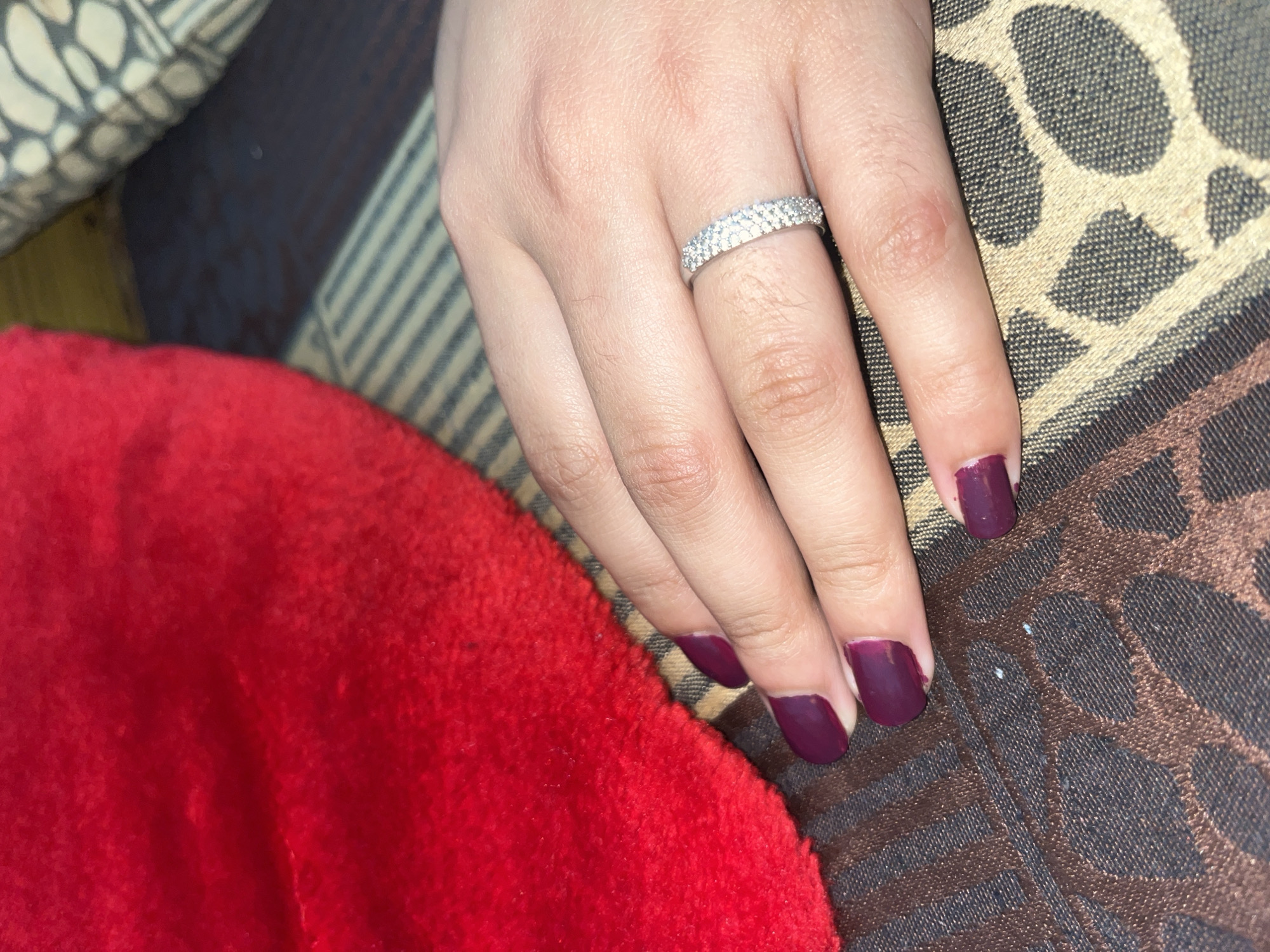 A close-up shot shows a person's left hand resting on a patterned fabric surface. The hand has fingernails painted with a deep plum color. A silver ring, encrusted with small, sparkling stones, is worn on the ring finger. To the left of the hand, a textured, bright red object is partially visible, likely made of a soft material like velvet or plush. The patterned fabric beneath the hand features a repeating design of dark brown and beige shapes. The lighting is moderate, casting soft shadows.