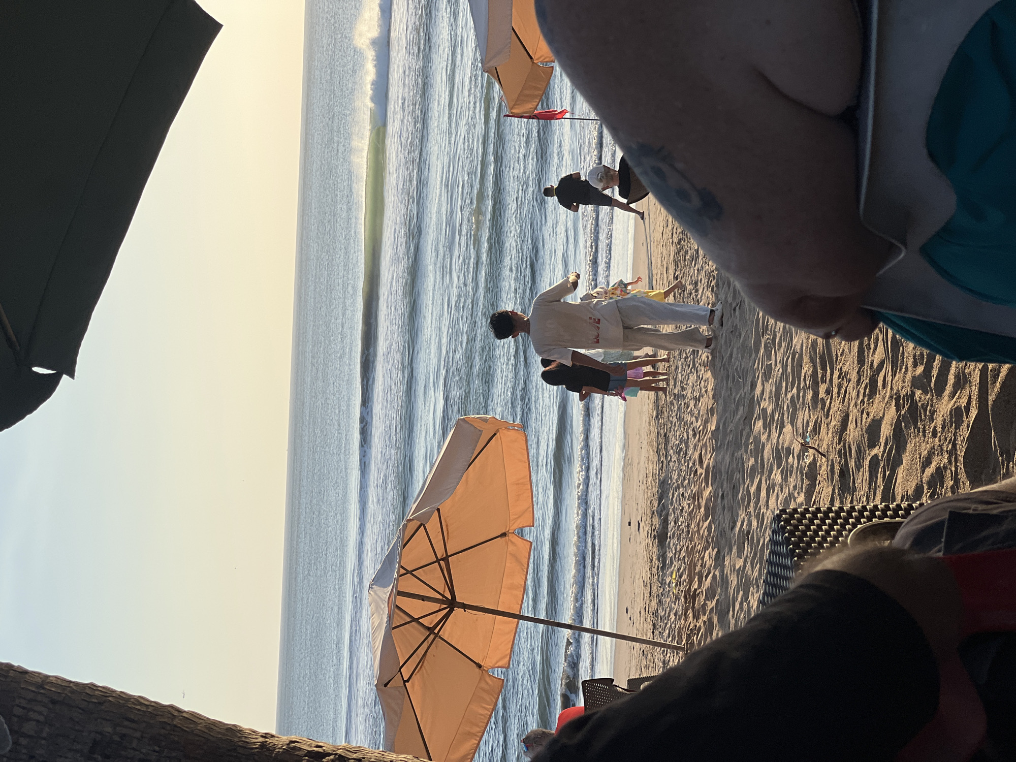 In Kuta, Indonesia, a serene beach scene unfolds at sunset. The golden hour bathes the sand and ocean in a warm glow. Several people are visible on the beach, some walking, others seated under umbrellas. In the foreground, the perspective is from someone seated, with their arm and shoulder visible, resting on a chair. A large orange beach umbrella dominates the left side of the frame, providing shade. Further back, other beach umbrellas and a red flag are visible. Gentle waves roll into the shore, with a surfer catching a wave in the distance. The sky is a soft gradient of blue and yellow, indicating the approaching dusk. The overall atmosphere is peaceful and relaxed, typical of a beach setting in Kuta.