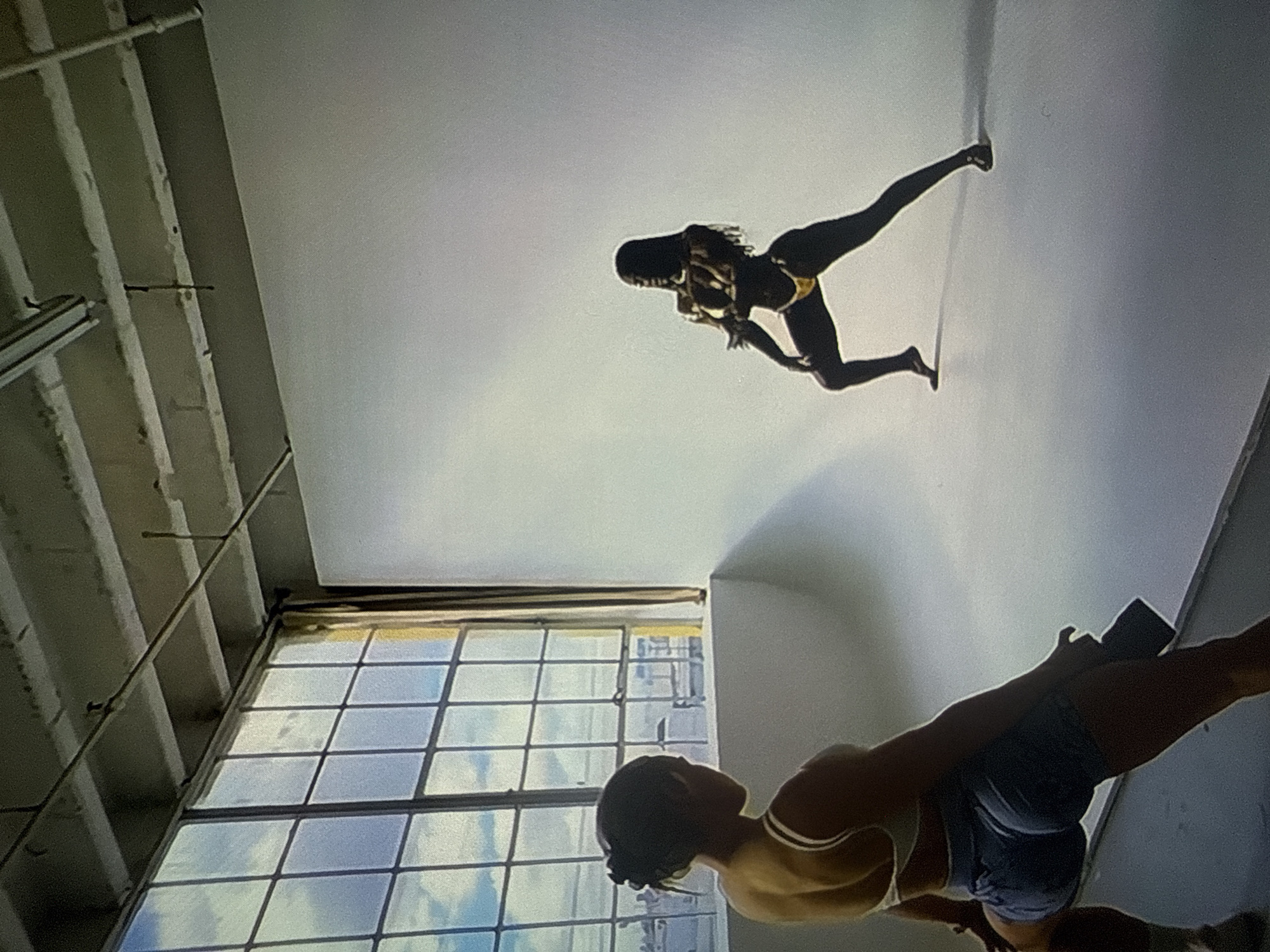 An indoor photograph captures a studio setting in Varna, Bulgaria. In the foreground, facing away from the viewer and towards the right, is an individual with dark hair tied back, wearing a light-colored sports bra and dark shorts. This individual holds a dark object, likely a camera, in their right hand, positioned as if actively photographing. In the mid-ground, on a seamless white backdrop that extends from the floor to the wall, stands a dark-skinned model wearing a yellow two-piece bikini. The model is striking a dynamic pose, with legs spread wide and arms bent, facing slightly right with a direct gaze. To the left of the scene, a large industrial-style window with multiple panes reveals a partially cloudy blue sky and distant urban structures. The ceiling features exposed concrete beams and visible utility piping. Shadows are cast on the white backdrop, indicating light primarily originates from the large window.