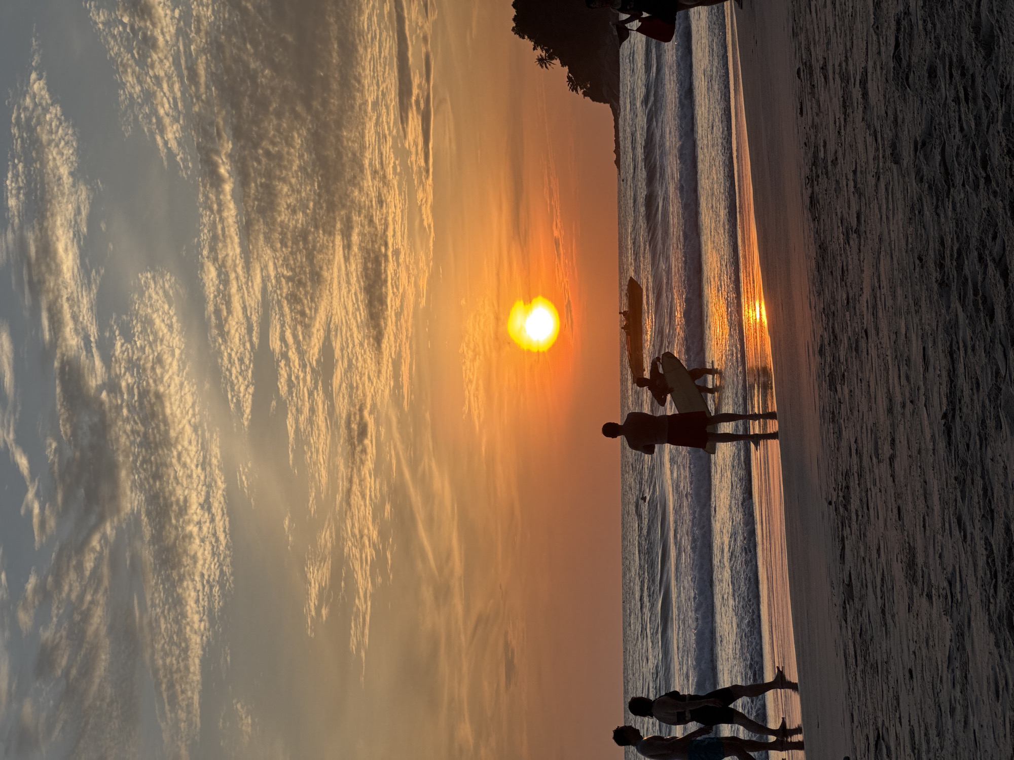 The image depicts a sunset scene at Karon Beach in Karon, Thailand. Silhouetted figures are visible on the sandy shore, some walking and others standing. One person is carrying a surfboard, suggesting recreational activities. The ocean features gentle waves, and a small boat with passengers is visible in the distance. The sky is filled with dramatic clouds, painted in warm hues of orange and yellow, indicating the sun is either rising or setting. The reflection of the sky is cast on the wet sand near the water's edge. A tree-covered headland is visible on the right side of the horizon, characteristic of tropical coastal landscapes.