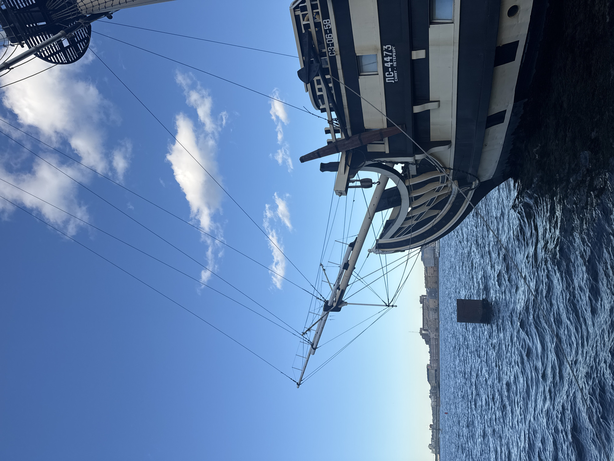 This is a low-angle, full-shot, exterior, daytime photograph of the stern section of a historical sailing ship docked in the Neva River, Saint Petersburg, Russia. The ship is painted black and white and has a classic, ornate design. The bowsprit extends to the left, pointing towards a distant cityscape that features buildings with varying architectural styles, including classical and Soviet-era structures. The Neva River is choppy with deep blue water. Several rigging lines stretch across the frame against a clear blue sky with scattered white clouds. A dark, rectangular metal buoy or marker protrudes from the water in the mid-ground, to the left of the ship's hull. The ship's hull bears Cyrillic inscriptions, including "ЛС-4473" and "САНКТ-ПЕТЕРБУРГ," indicating its identification and location. Another inscription, "СЗ-06-58," is visible higher up on the ship's structure. The overall scene conveys a sense of historical maritime presence within the urban landscape of Saint Petersburg. There are no people visible in the frame.
