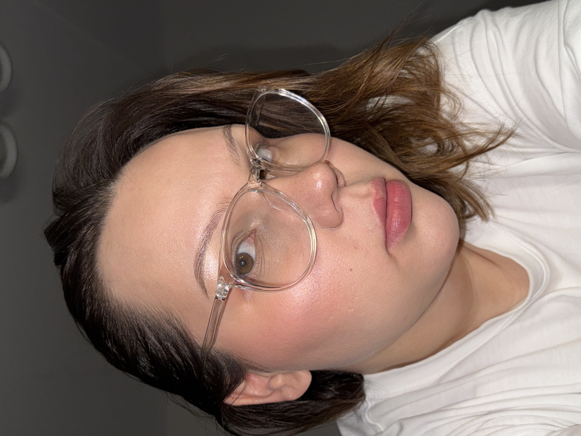A close-up, angled, selfie-style photograph depicts a fair-skinned woman with dark brown hair featuring lighter brown highlights, wearing clear-framed glasses. The woman is wearing a white t-shirt, and her face is angled towards the camera. Her lips are a natural pink color. The background is a muted gray tone, with a blurred white object visible in the upper left corner, likely a light fixture. No specific indicators point to the location being Chaikovsky, Russia; no buildings, landmarks, or signage are visible.
