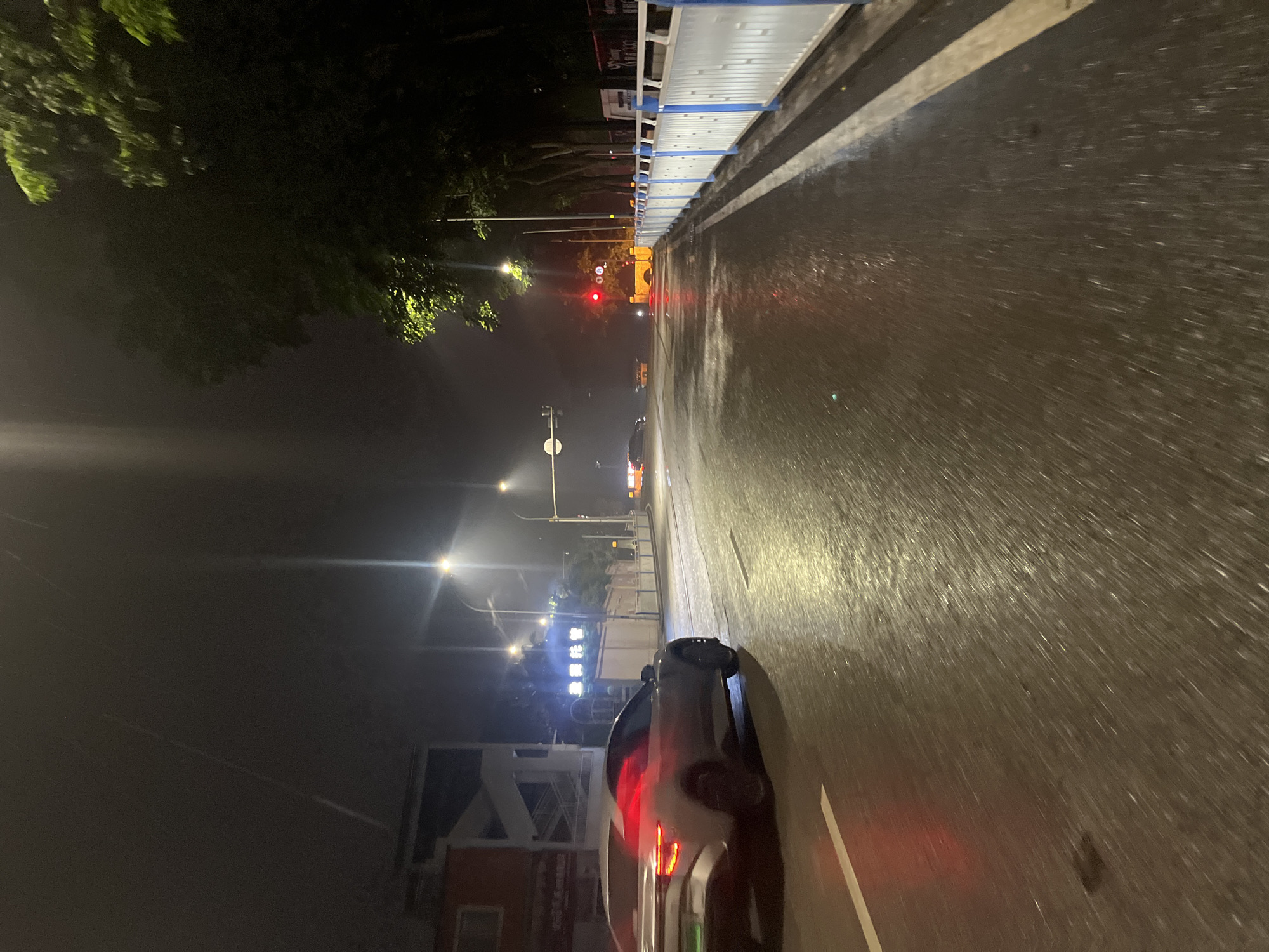 The image depicts a nighttime scene on a wet street in Jiulongpo District, China. A silver sedan with its red taillights illuminated is driving on the right side of the road. To the left of the road, there's a building with some windows and an external staircase. Streetlights are visible, casting a glow on the wet asphalt. Further down the road, a traffic light shows red, and other vehicles are present in the distance. Trees line the right side of the road, and a white barrier with blue accents runs along the edge of the sidewalk. The atmosphere appears somewhat hazy, possibly due to rain or fog, which is reflected in the wet road surface.