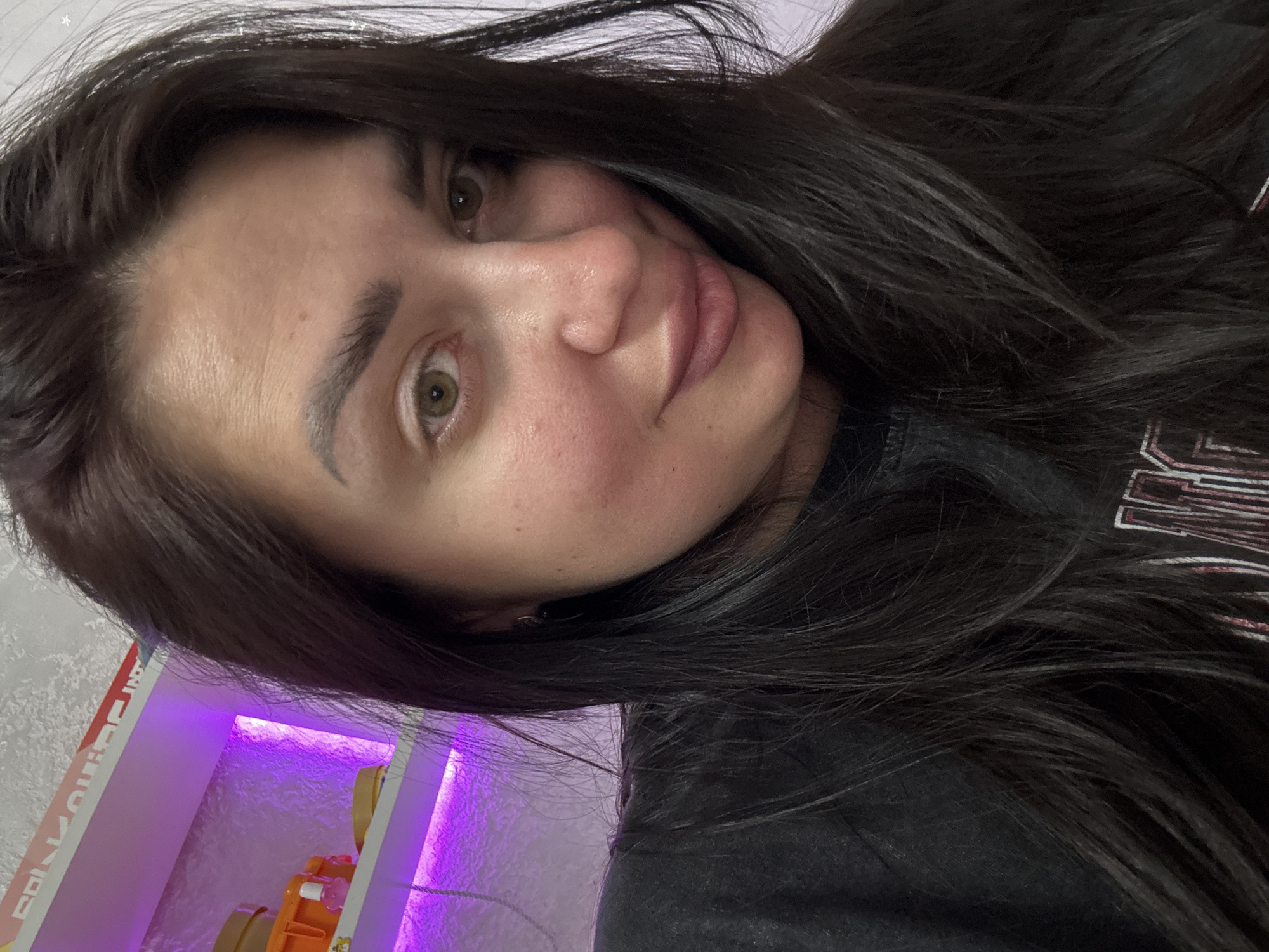 The image is a close-up selfie of a woman with dark hair. She is wearing a black t-shirt and has light brown eyes. Her skin appears to have a rosy tone. In the background, there is a shelf with purple lighting, displaying several containers and a small bottle. The overall environment suggests an indoor setting, possibly a home or a personal space. The location in Tolyatti, Russia, is not discernible from the visual information provided in the image.