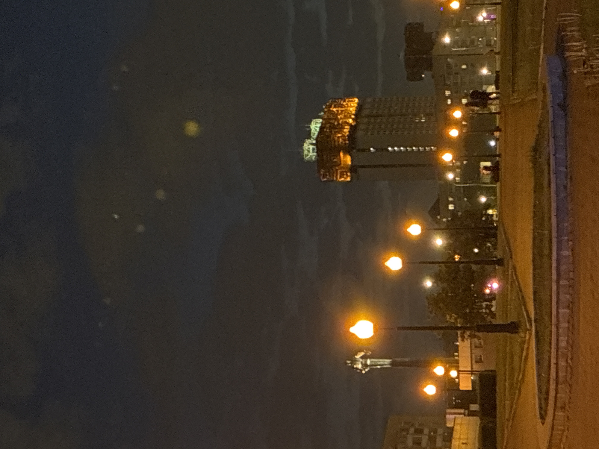 The image depicts an urban night scene in Moscow, Russia. A row of illuminated street lamps lines a walkway. Buildings, including a prominent skyscraper with golden lights, are visible in the background. There are a few people walking along the walkway. The sky is dark and cloudy.