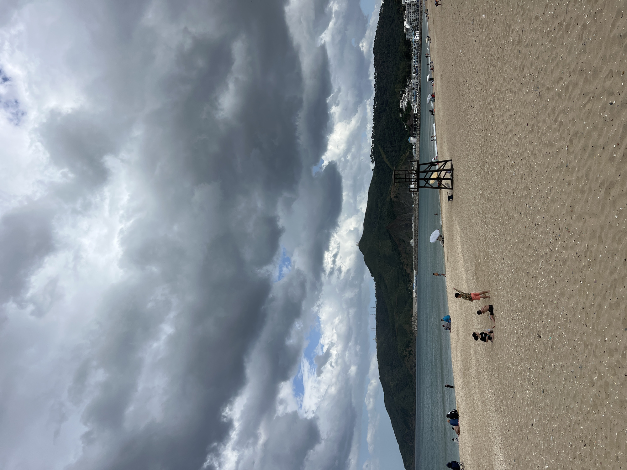 The image depicts a beach in M'Diq, Morocco. A mountain range covered in vegetation runs along the coast, with buildings visible at its base. A lifeguard stand sits on the sandy beach. Several people are scattered along the beach, some appearing to be seated or bending over, while others are standing. The sky is filled with heavy, gray clouds, suggesting an overcast day. The sea is calm.