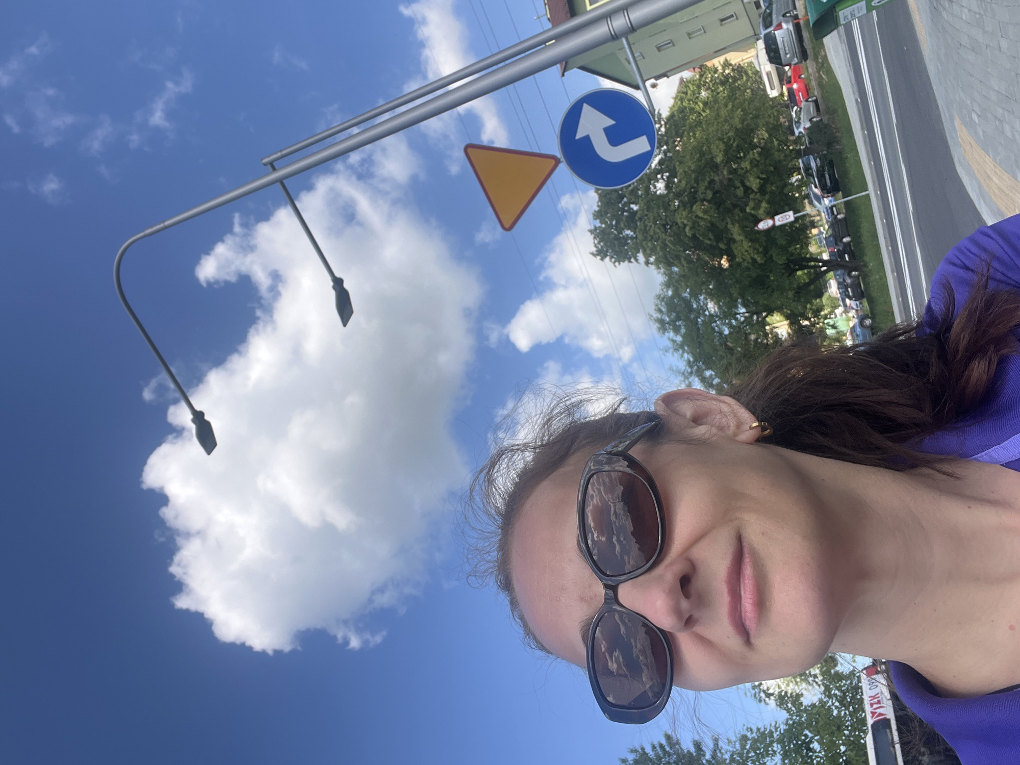 A low-angle, waist-up shot features a woman with brown hair and sunglasses, looking up towards the sky. She is wearing a purple polo shirt. The background consists of a bright blue sky with fluffy white clouds, streetlights, and traffic signs. A yellow triangular sign and a blue circular sign with a white arrow indicating a right turn are visible. Trees and buildings are present in the background, along with parked cars and a street. A portion of a bus is visible on the far left. The scene appears to be an urban environment on a sunny day.