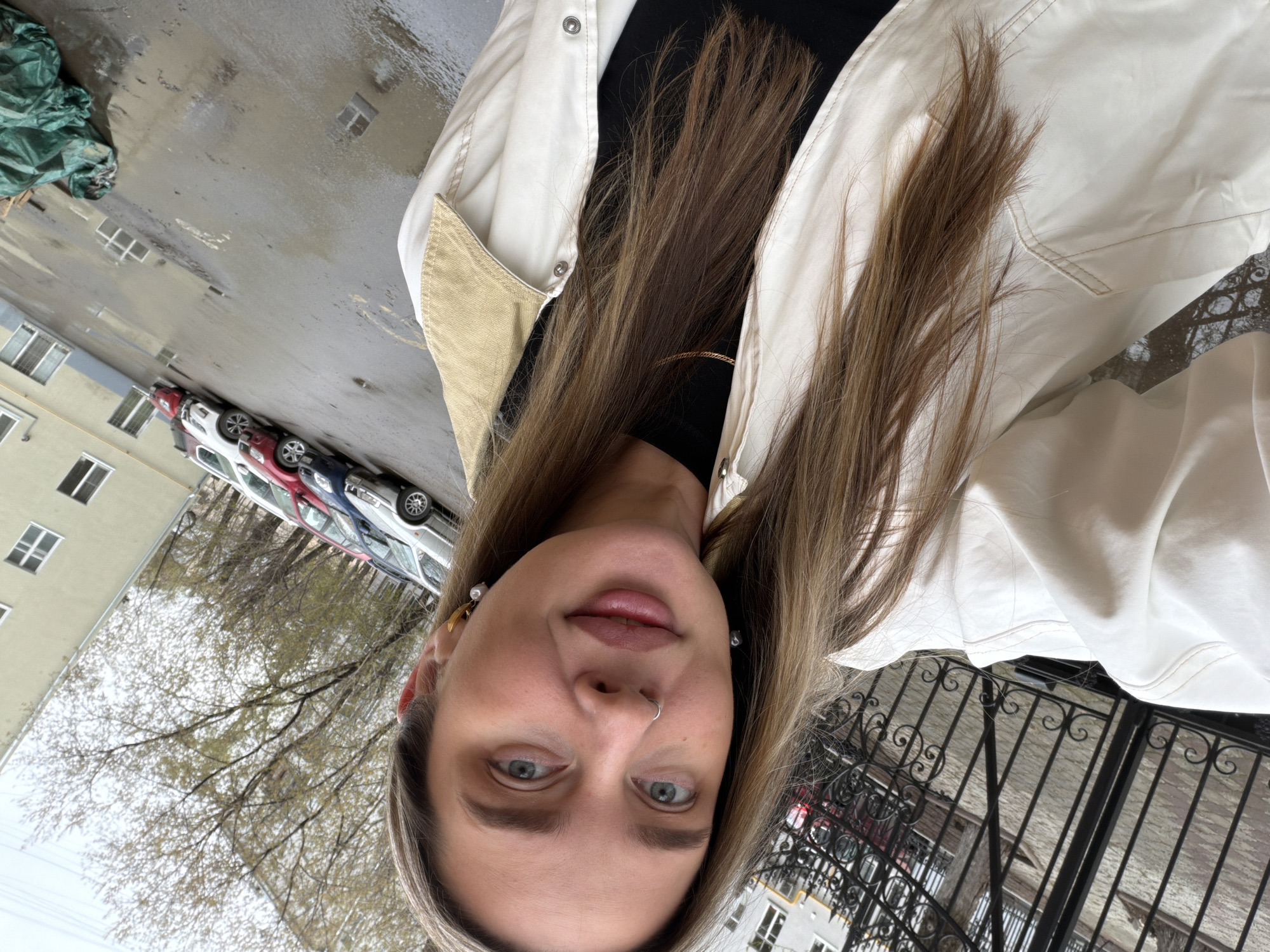 A portrait-style image, taken outdoors in what appears to be Moscow, Russia, features a person, likely a woman, captured from the chest up. The individual has long, straight brown hair, blue eyes, and a subtle smile. They are wearing a white collared jacket with beige or tan accents on the shoulders, over a black top. A silver hoop nose ring is visible, along with earrings: one is a gold-toned crescent shape with a pearl, and the other appears to be a pearl stud.
The background suggests an urban or suburban environment. The ground is paved and wet, indicating recent rain, with reflections of buildings and structures visible. To the left, a light-colored building with multiple rectangular windows stands alongside a row of parked cars, including red, white, and dark-colored vehicles. A green tarp or covering lies on the wet ground. Bare tree branches or trees with very early spring foliage are visible against an overcast sky. On the right side of the image, a decorative black wrought-iron fence or gate with swirling patterns is partially visible. The scene depicts a daytime setting with cool, overcast weather conditions. There is no visible text in the image.