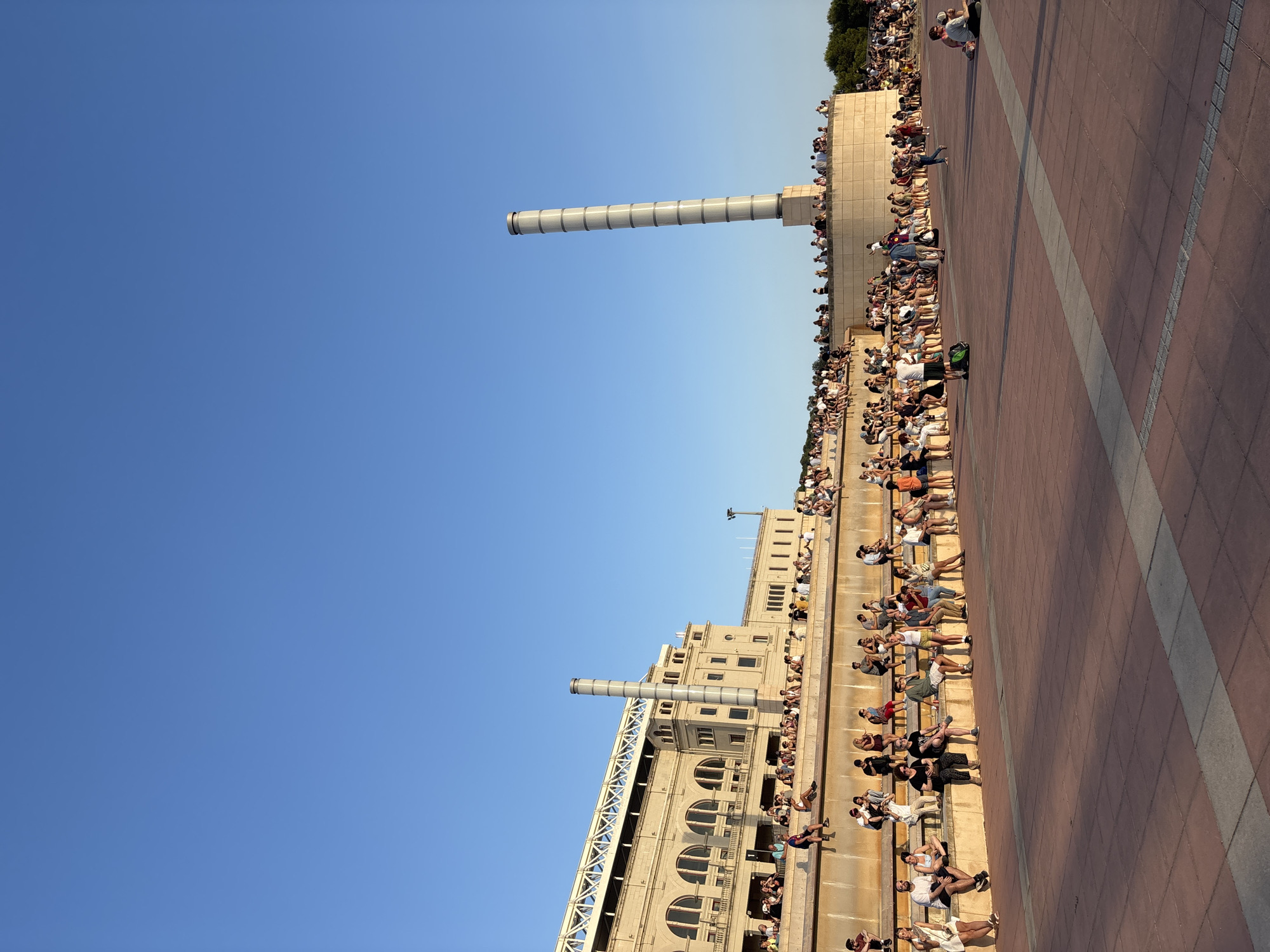 The image depicts a wide-angle view of the Olympic Ring area of Barcelona, Spain. The scene is bathed in the warm light of late afternoon, with a clear blue sky dominating the upper portion of the frame. In the background, the iconic Palau Nacional, a grand neoclassical building, stands with its distinctive arched windows and classical facade. To its right, a tall, cylindrical tower with segmented grey bands, part of the Olympic Ring's architecture, rises into the sky.
In the mid-ground, a tiered seating area is filled with a diverse crowd of people. They are seated and standing on the stone steps, facing outwards towards an open plaza. The people are dressed casually in summer attire, engaging in various activities such as conversing, relaxing, and observing the surroundings. Some are sitting individually, while others are gathered in small groups.
The foreground consists of a vast, red-tiled plaza, marked by lighter-colored stripes. The plaza is largely empty except for a few individuals scattered across its expanse. One person is seen walking, and another is seated on the ground. The overall impression is of a popular public space on a pleasant day, with many people enjoying the open area and the architectural features of the Olympic Ring. The sunlight casts long shadows across the plaza, indicating the time of day.